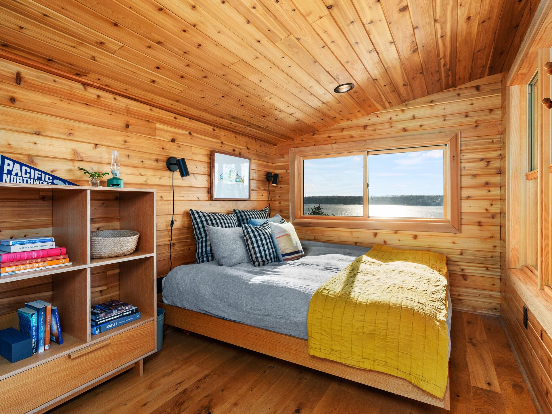 A cozy wooden room features a bed with gray and yellow bedding, a bookshelf filled with books, and a large window offering a scenic view of a lake. The walls and ceiling are crafted from wood, creating a warm, rustic ambiance.