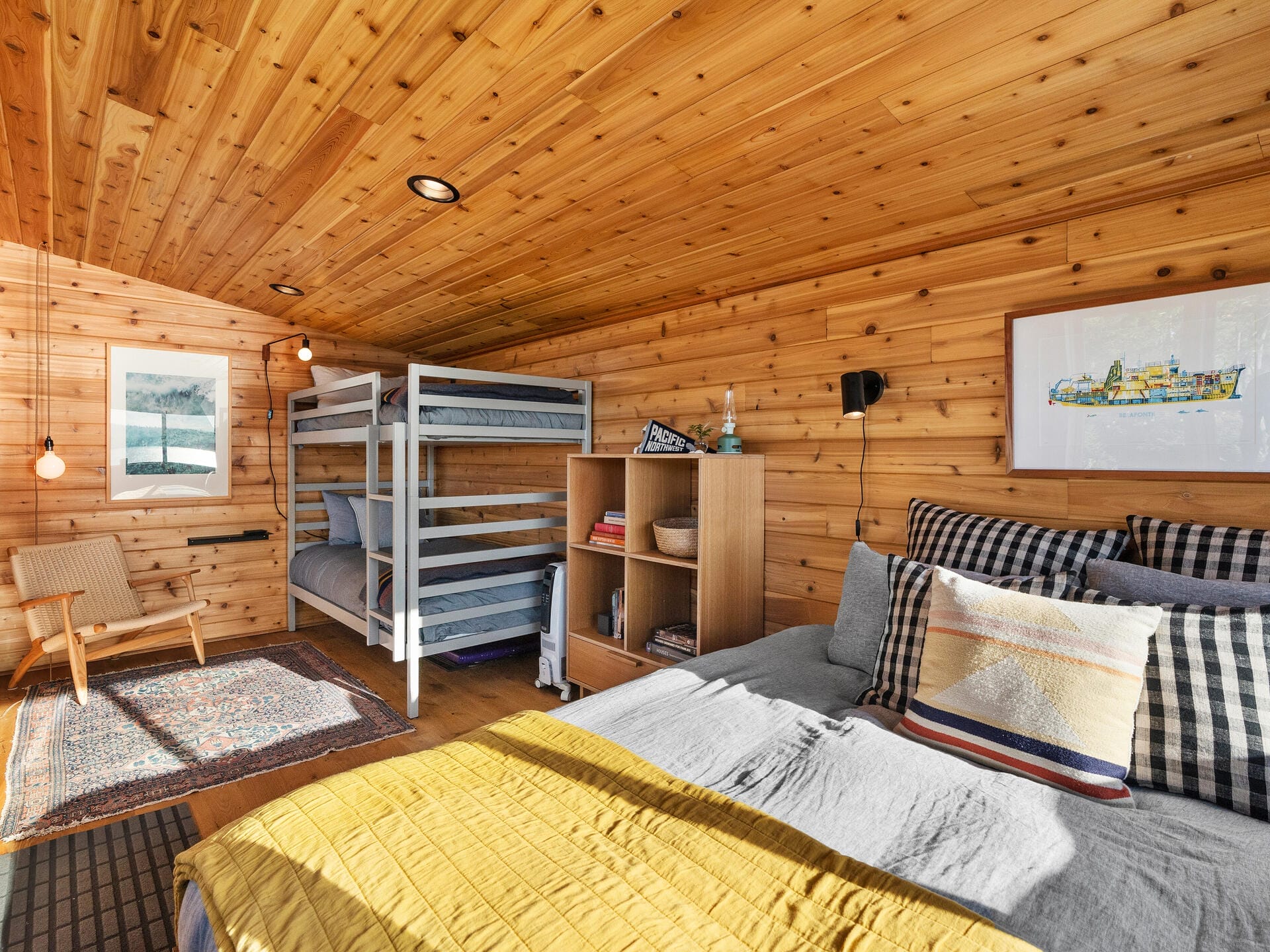 A cozy room with wooden walls and ceiling features a bunk bed, a queen-sized bed with colorful pillows, a wooden chair, and a small bookshelf. Two framed pictures hang on the walls, and a warm rug lies on the floor. Soft lighting adds warmth.