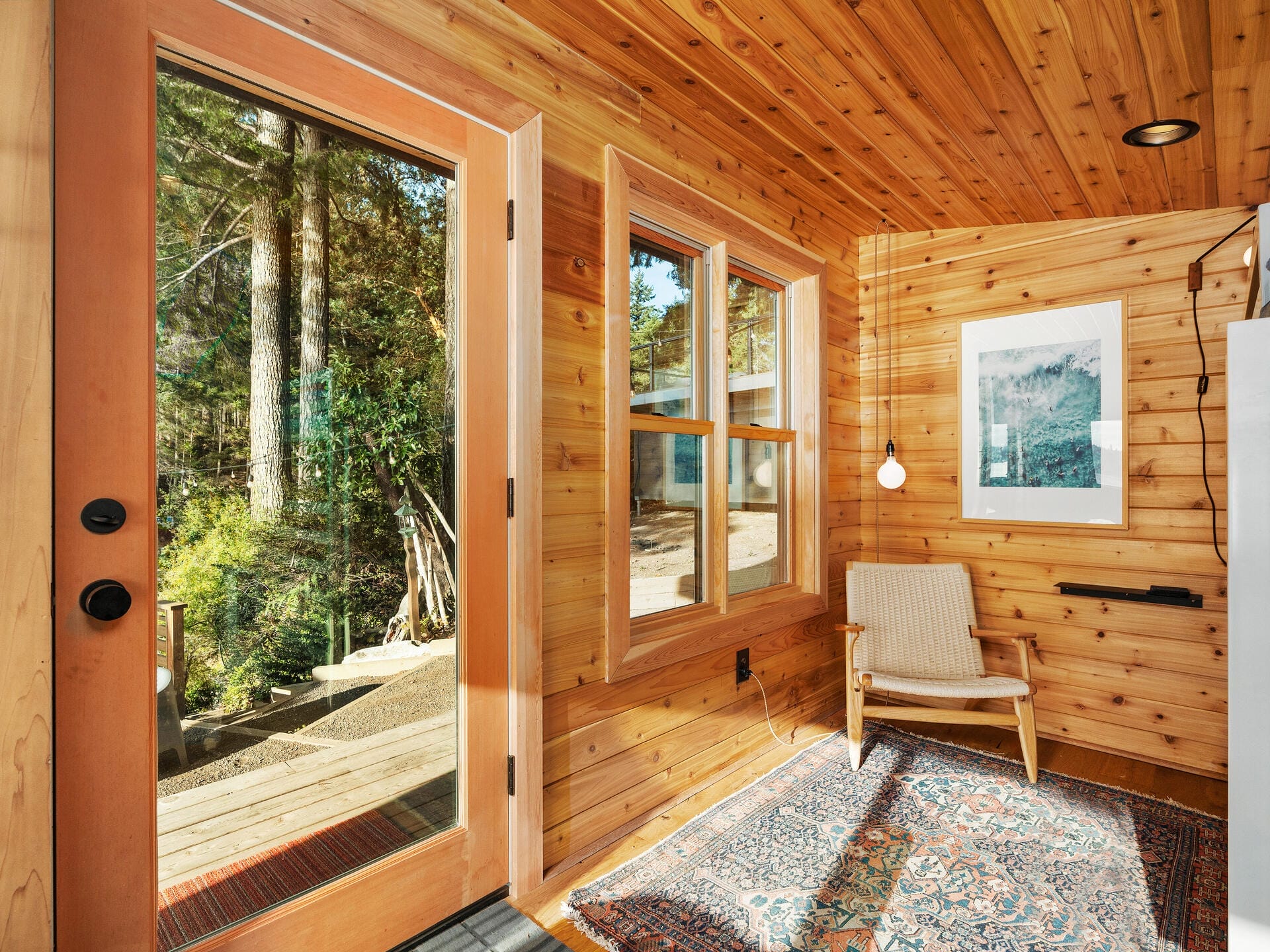 A cozy room with wooden walls and ceiling, featuring a large window and glass door that open to a view of trees. Inside, there is a patterned rug, a white cushioned chair, a small pendant light, and framed artwork on the wall.