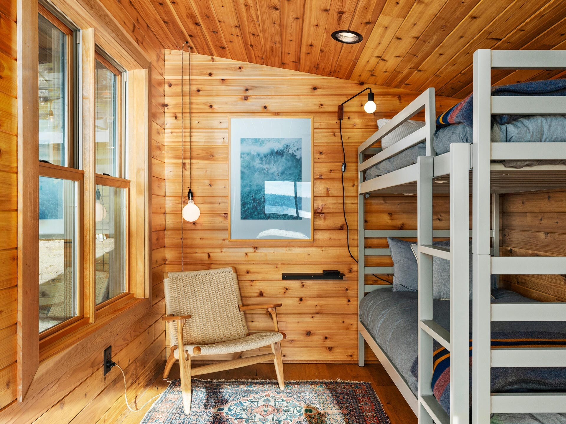 A cozy wooden room features a bunk bed with gray bedding, a woven chair, a patterned rug, and a framed mountain picture on the wall. Two round pendant lights hang from the ceiling, and a large window lets in natural light.