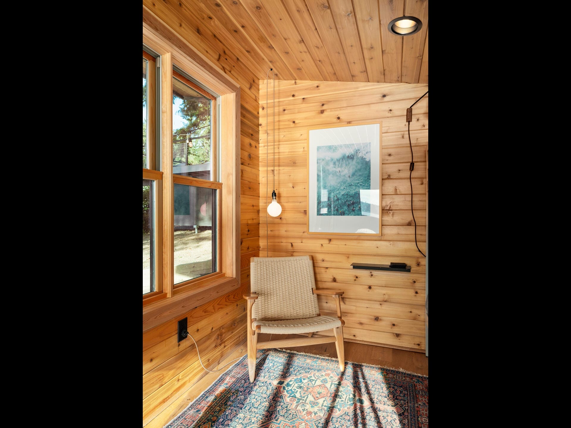 A cozy wooden room with a large window, armchair, and small hanging light. A framed picture hangs on the wall, and a colorful rug covers part of the floor. Sunlight streams in, creating a warm atmosphere.