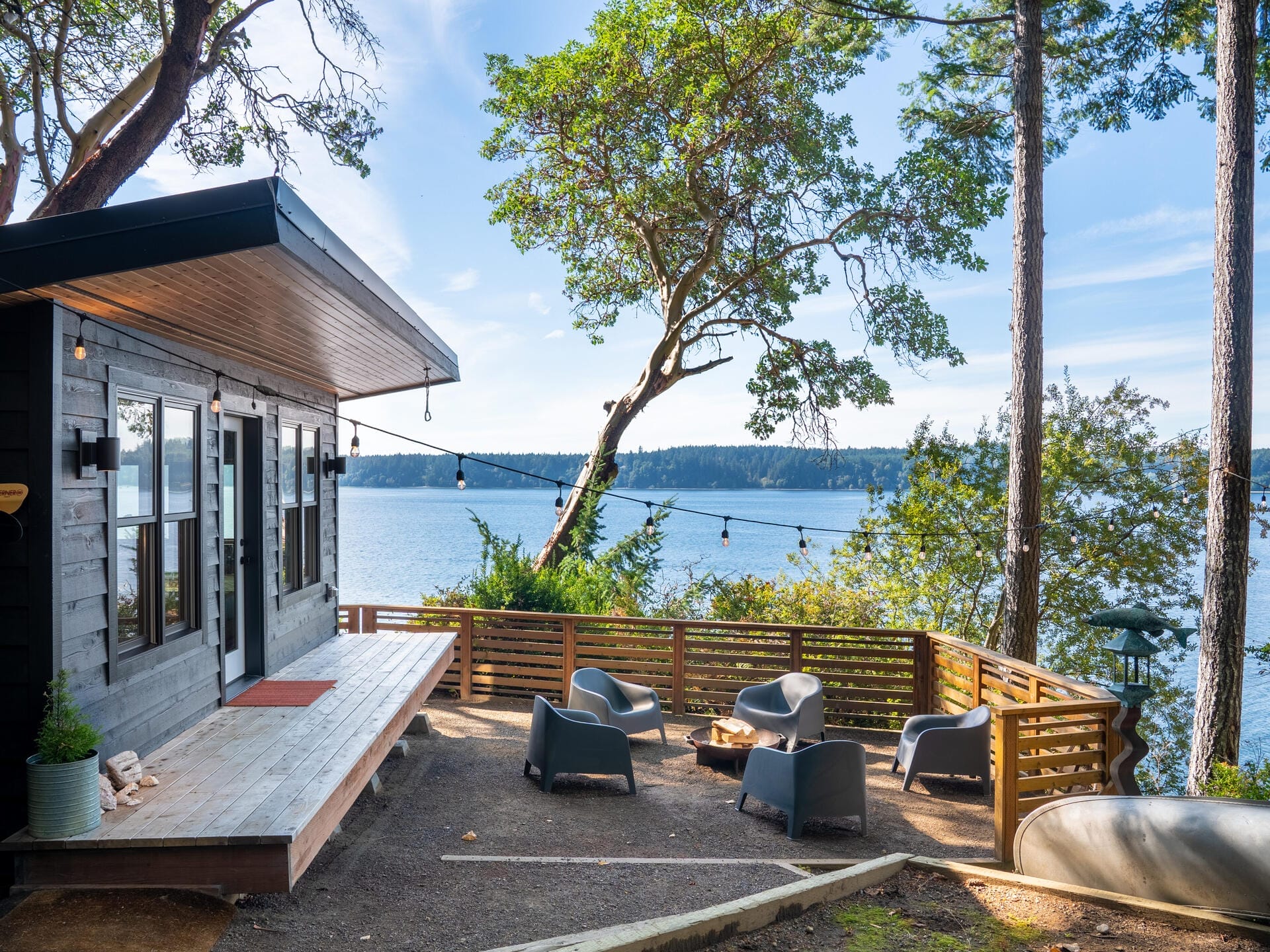 A modern cabin with large windows overlooks a serene lake. The deck features string lights and a seating area with grey chairs around a fire pit. Tall trees frame the view, and a canoe is visible nearby.