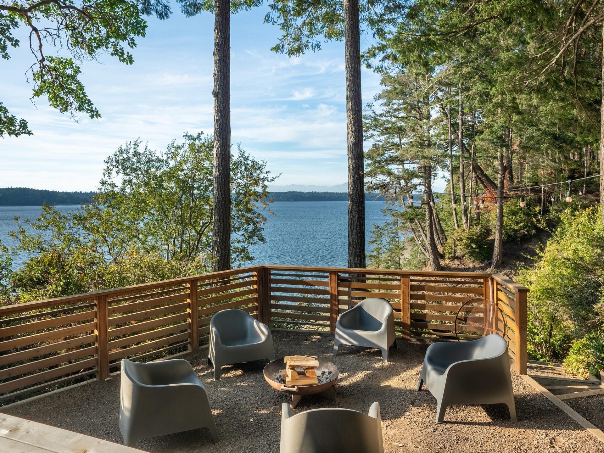 A serene outdoor seating area overlooking a calm body of water, surrounded by tall trees. The space includes modern chairs around a circular wooden table on a gravel patio, enclosed by a wooden railing.