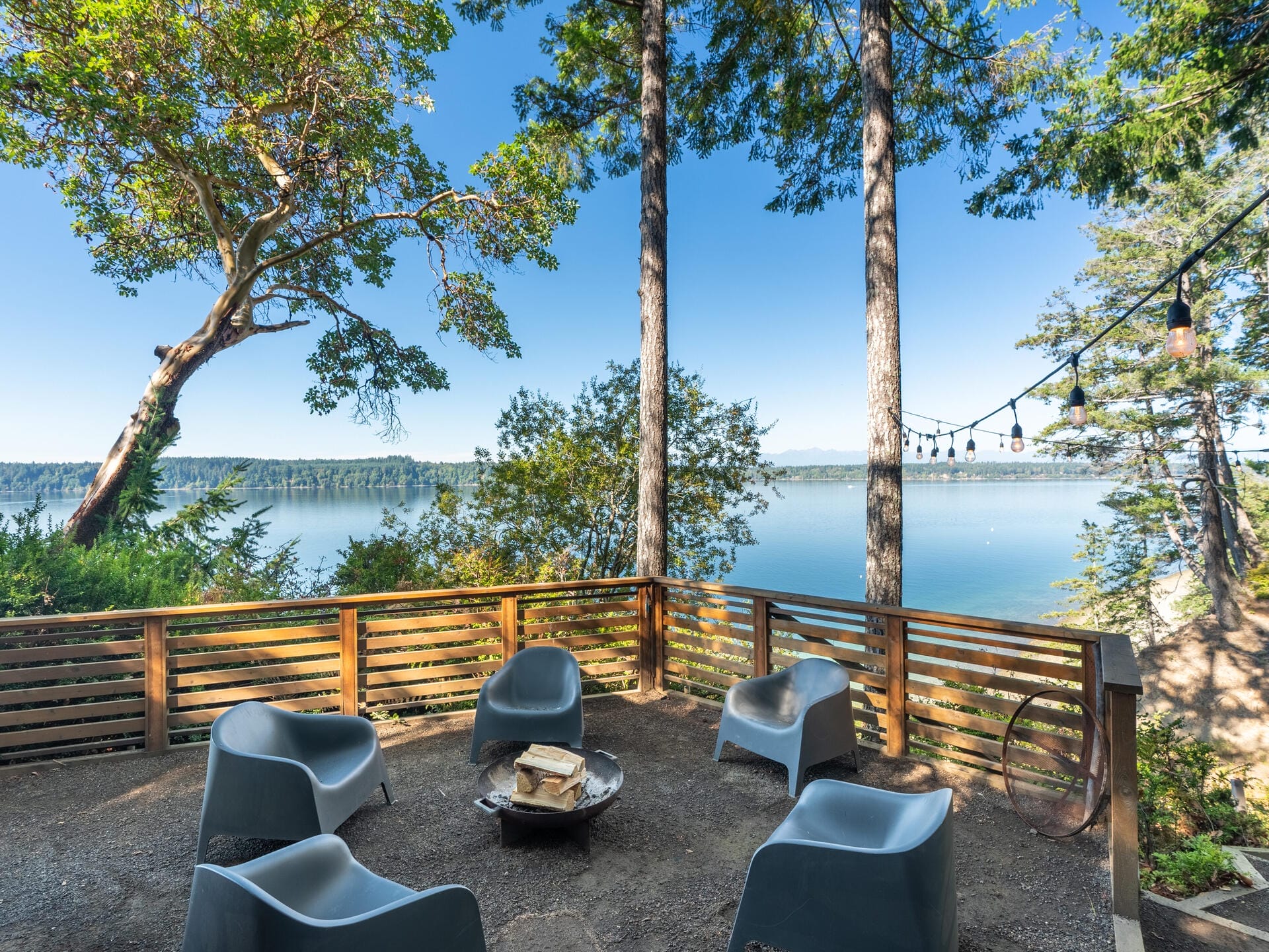 A serene lakeside view featuring a wooden patio with modern gray chairs surrounding a small table. String lights hang above, and tall trees frame the clear blue sky and water in the background, creating a tranquil outdoor setting.