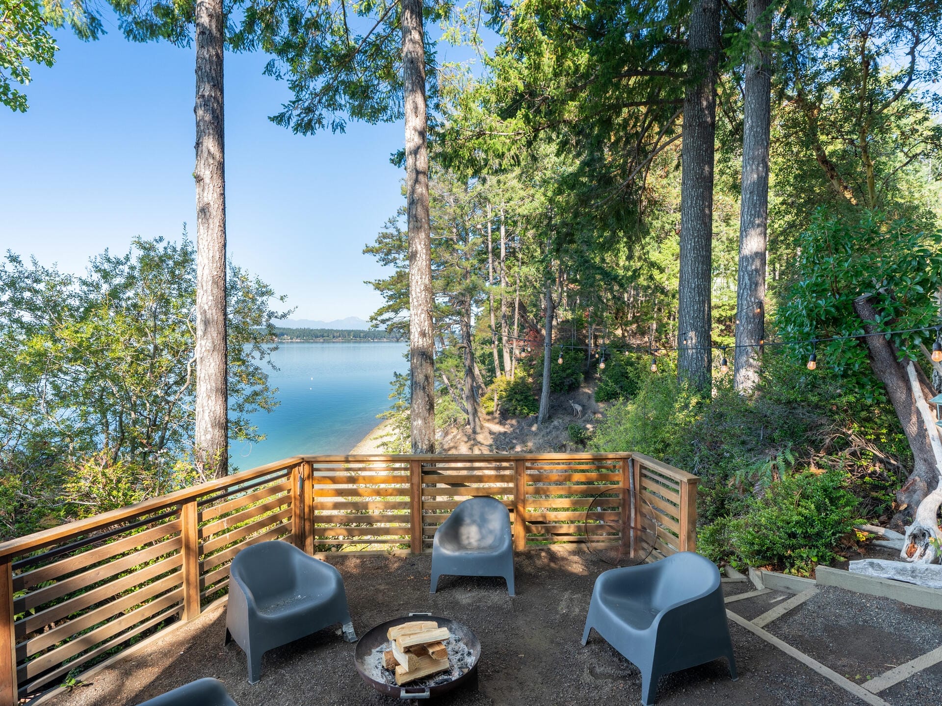 Outdoor seating area with gray chairs encircling a small fire pit on a wooden deck. Surrounded by tall trees, the deck overlooks a scenic view of a calm lake and distant hills under a clear blue sky.