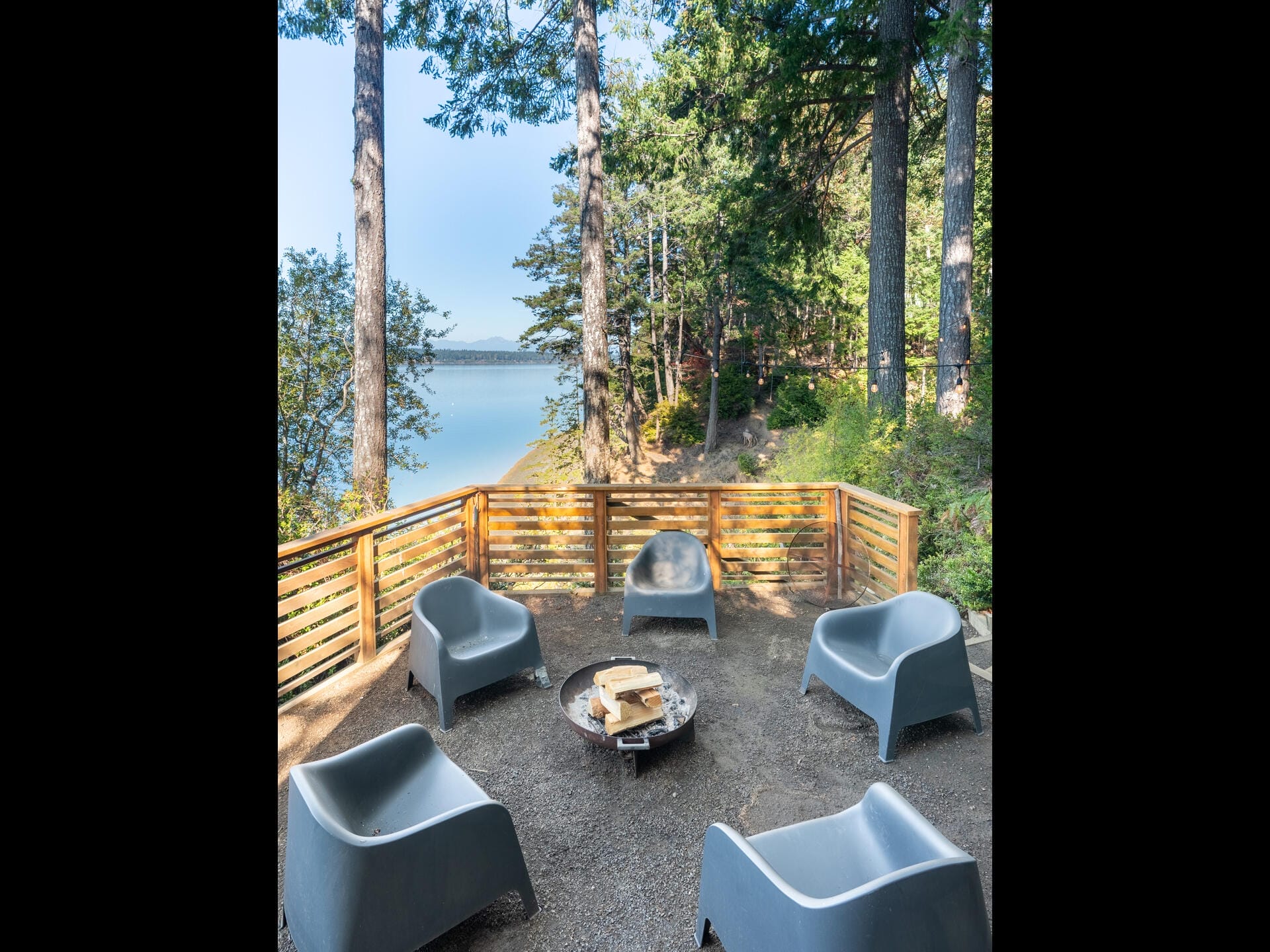 Outdoor seating area with modern gray chairs around a fire pit on a gravel patio, surrounded by trees. A wooden railing overlooks a serene lake under a clear blue sky.