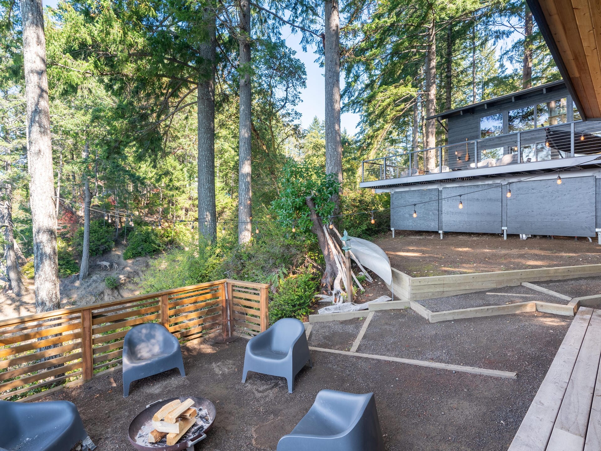 A backyard with modern gray chairs around a fire pit, overlooking a wooded area. A contemporary house with a deck and string lights is visible in the background, surrounded by tall trees.