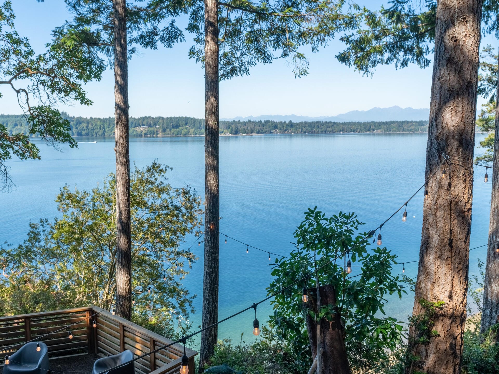 A serene lakeside view with calm blue waters bordered by lush green trees. String lights are hung between the trees, and a wooden deck is visible in the foreground. Mountains can be seen in the distant background under a clear blue sky.