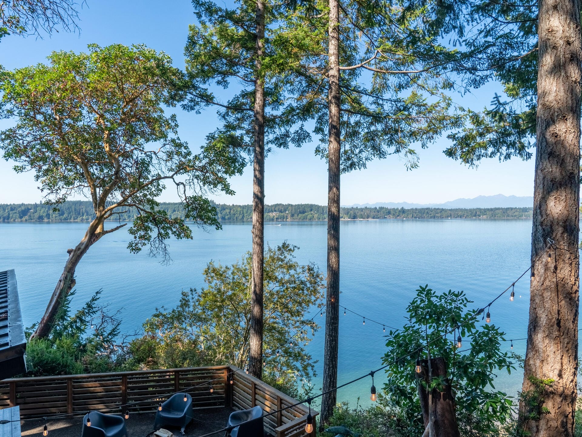 View of a serene lake surrounded by lush trees under a clear blue sky. A wooden deck with chairs overlooks the water, and string lights hang between the trees. Mountains are visible in the distance across the lake.