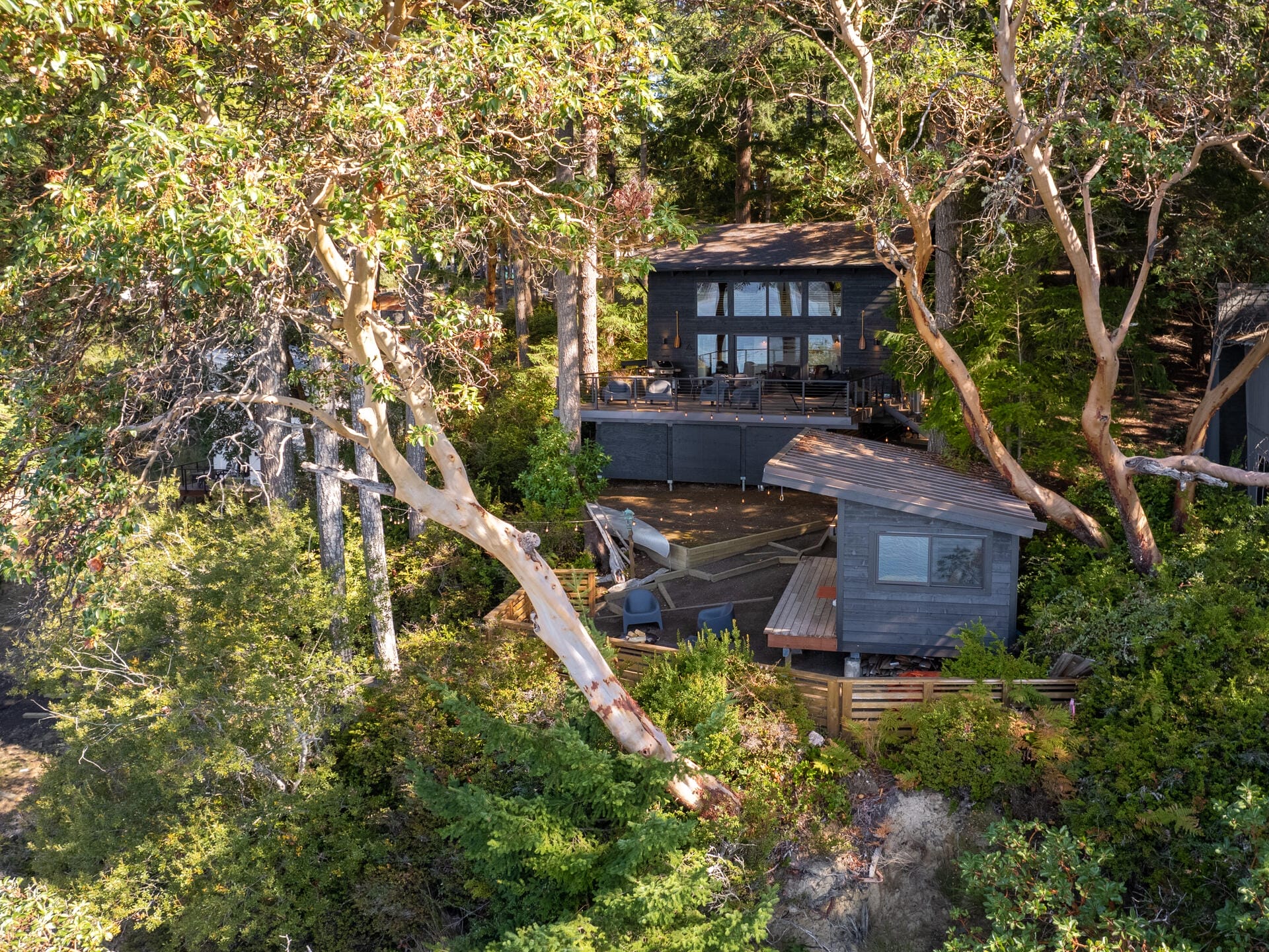 A modern black cabin with large windows nestled among trees on a hillside. The cabin features two levels, with an outdoor deck and a smaller outbuilding nearby. The surrounding area is lush with greenery and natural vegetation.