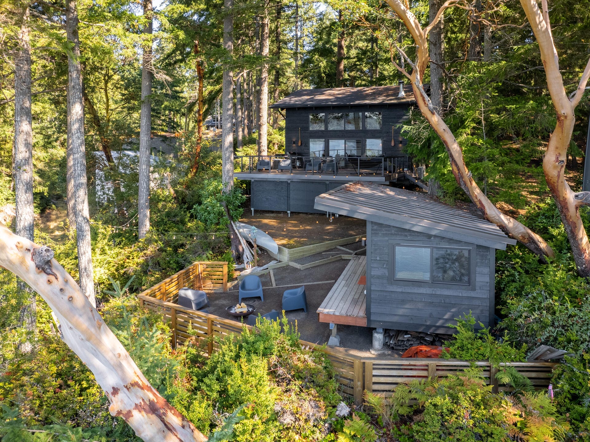 A cozy cabin nestled in a lush forest, featuring two dark gray structures with large windows. The outdoor space includes seating and a surfboard, surrounded by dense greenery and tall trees.