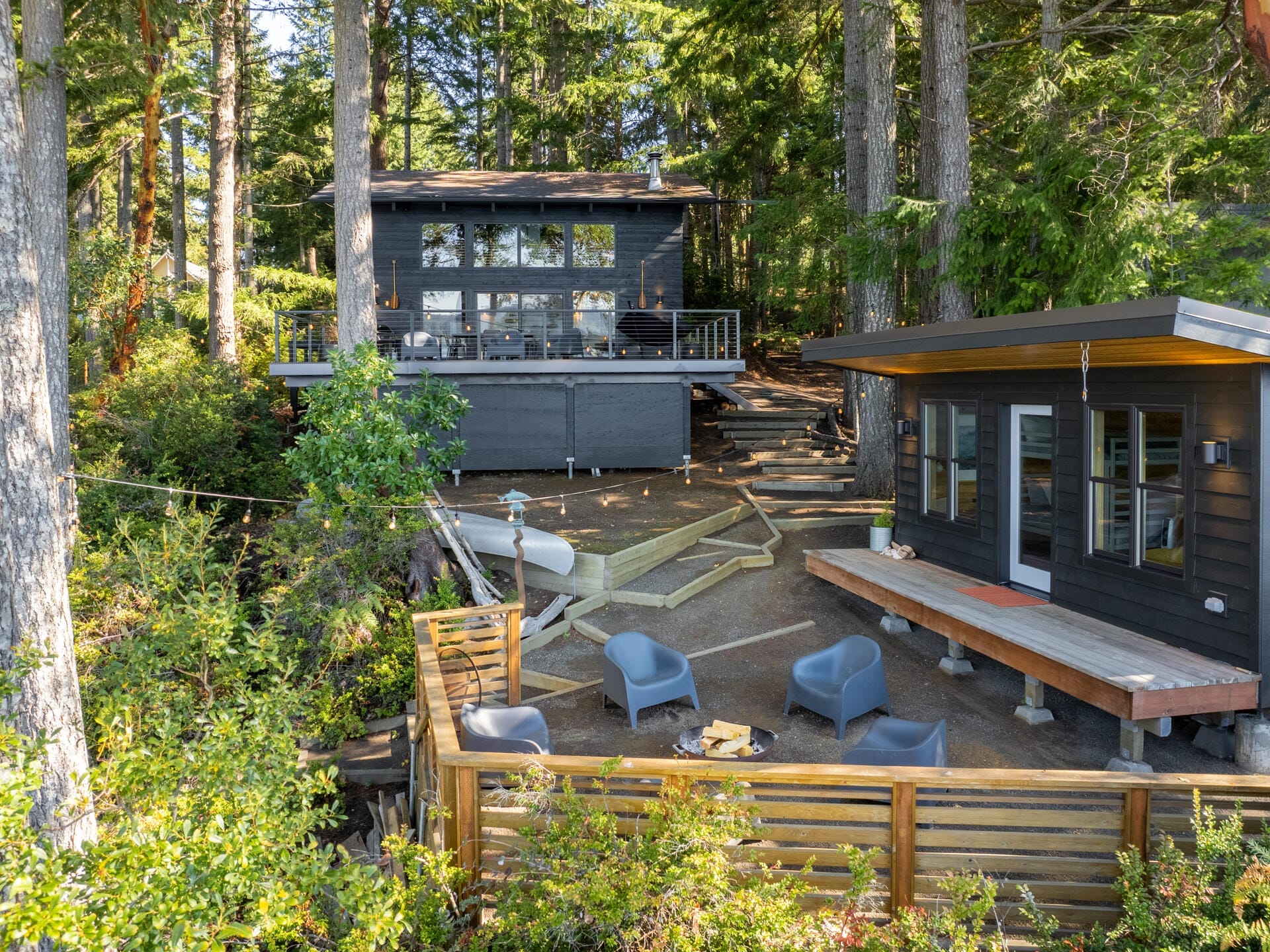 A cozy forest retreat with two modern black cabins surrounded by tall trees. The area features a fire pit with seating and a wooden deck overlooking the tranquil forest setting. A boat is docked nearby, adding to the serene atmosphere.