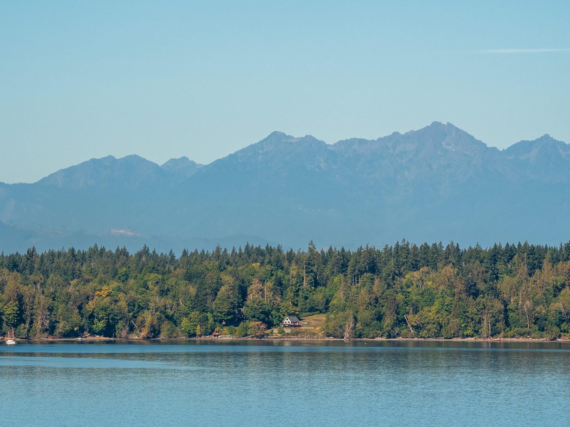 A serene landscape of a calm lake in the foreground, a forested area along the shoreline, and a backdrop of majestic, hazy mountains under a clear blue sky.