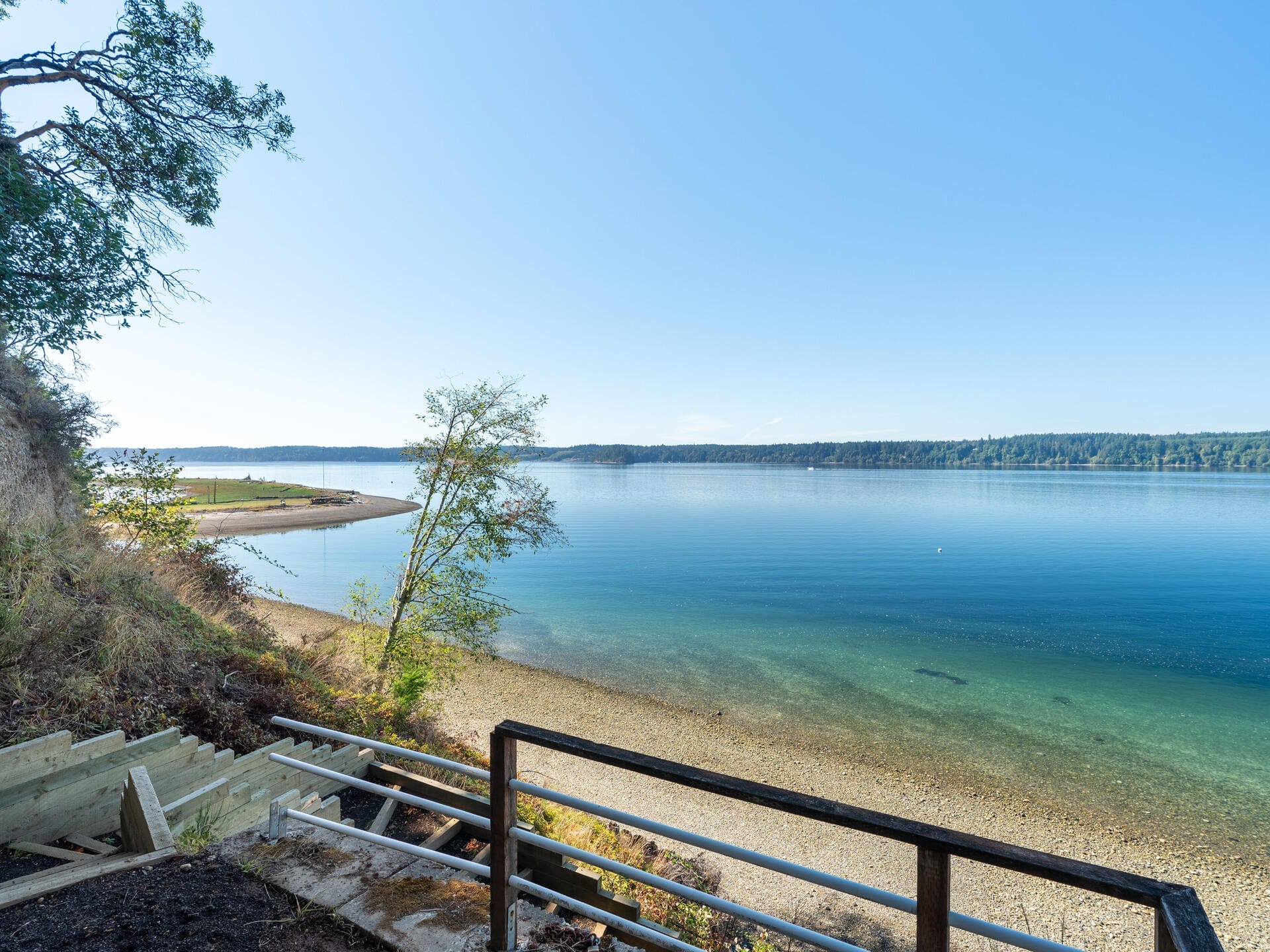 A tranquil lake scene with clear blue water extends to the distant tree-lined shore. A small pebbled beach and wooden stairs leading down to the water are visible in the foreground, with a clear blue sky overhead.