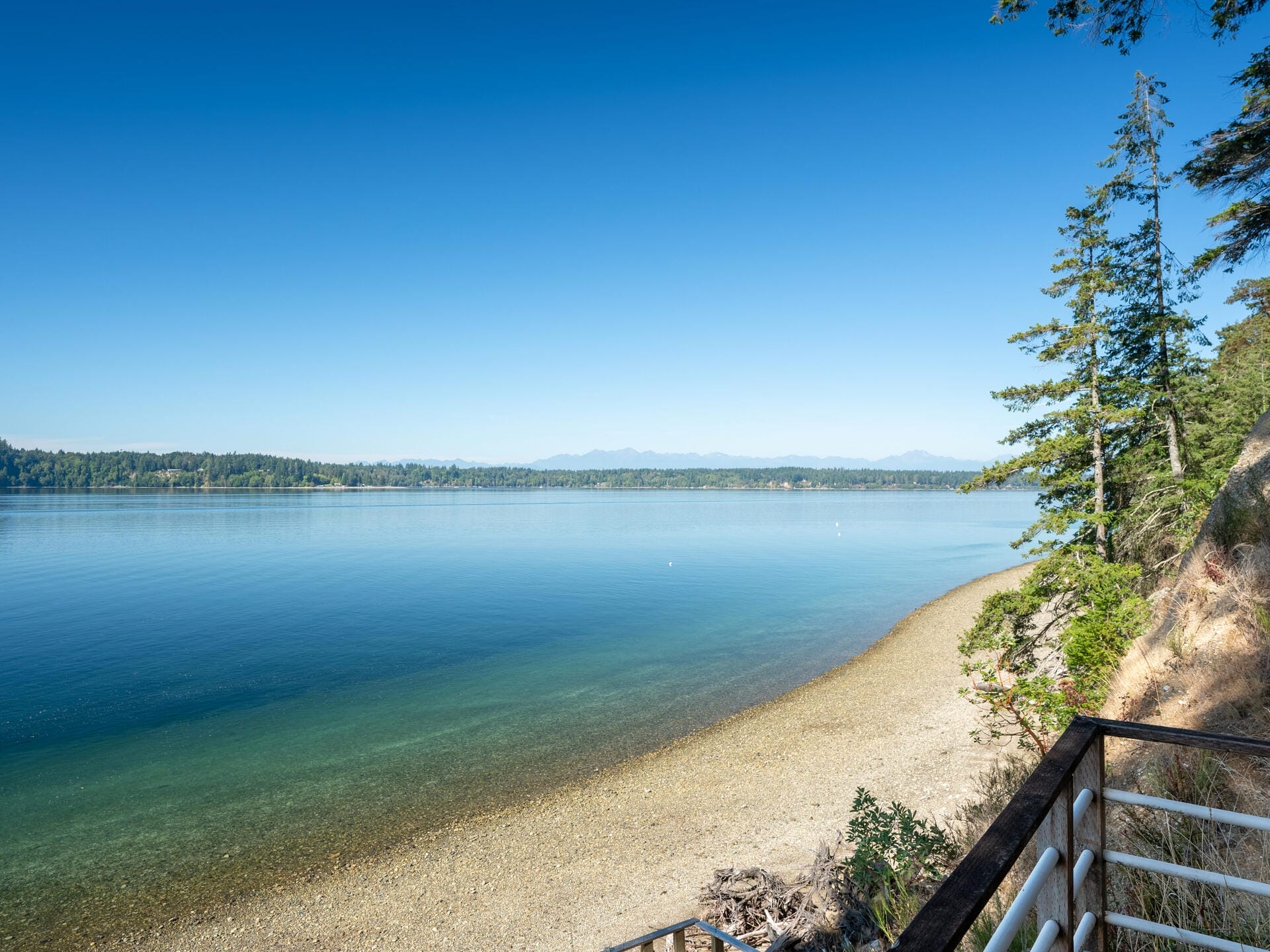 A scenic view of a calm, clear lake with a pebbled shoreline under a bright blue sky. Pine trees line the right side, and distant mountains are visible on the horizon. The foreground features a railing overlooking the water.