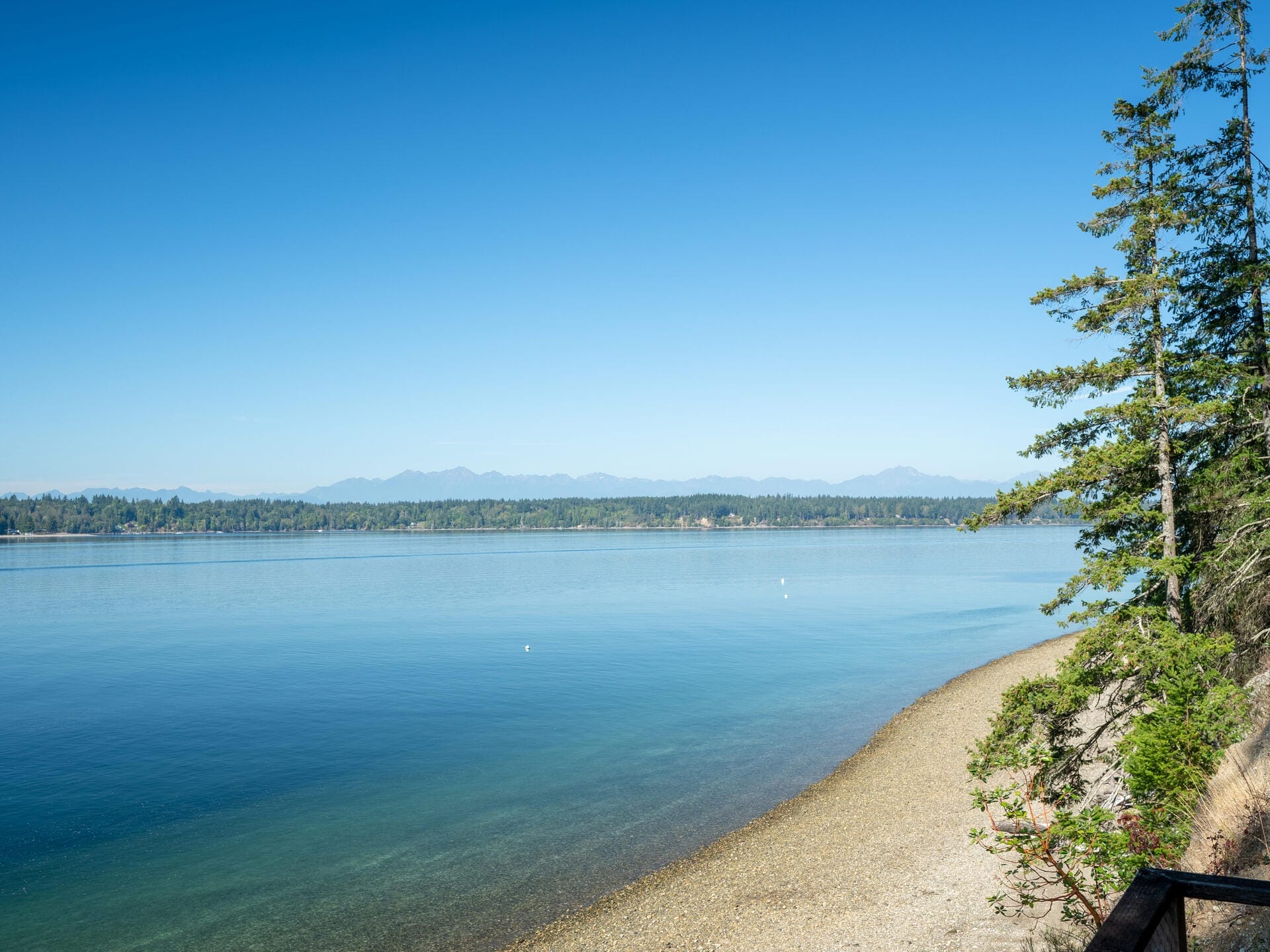 A serene lakeside scene with clear blue water reflecting the sky, a pebbled shoreline, and distant evergreen trees under a clear, bright sky. Mountain peaks are visible on the horizon.