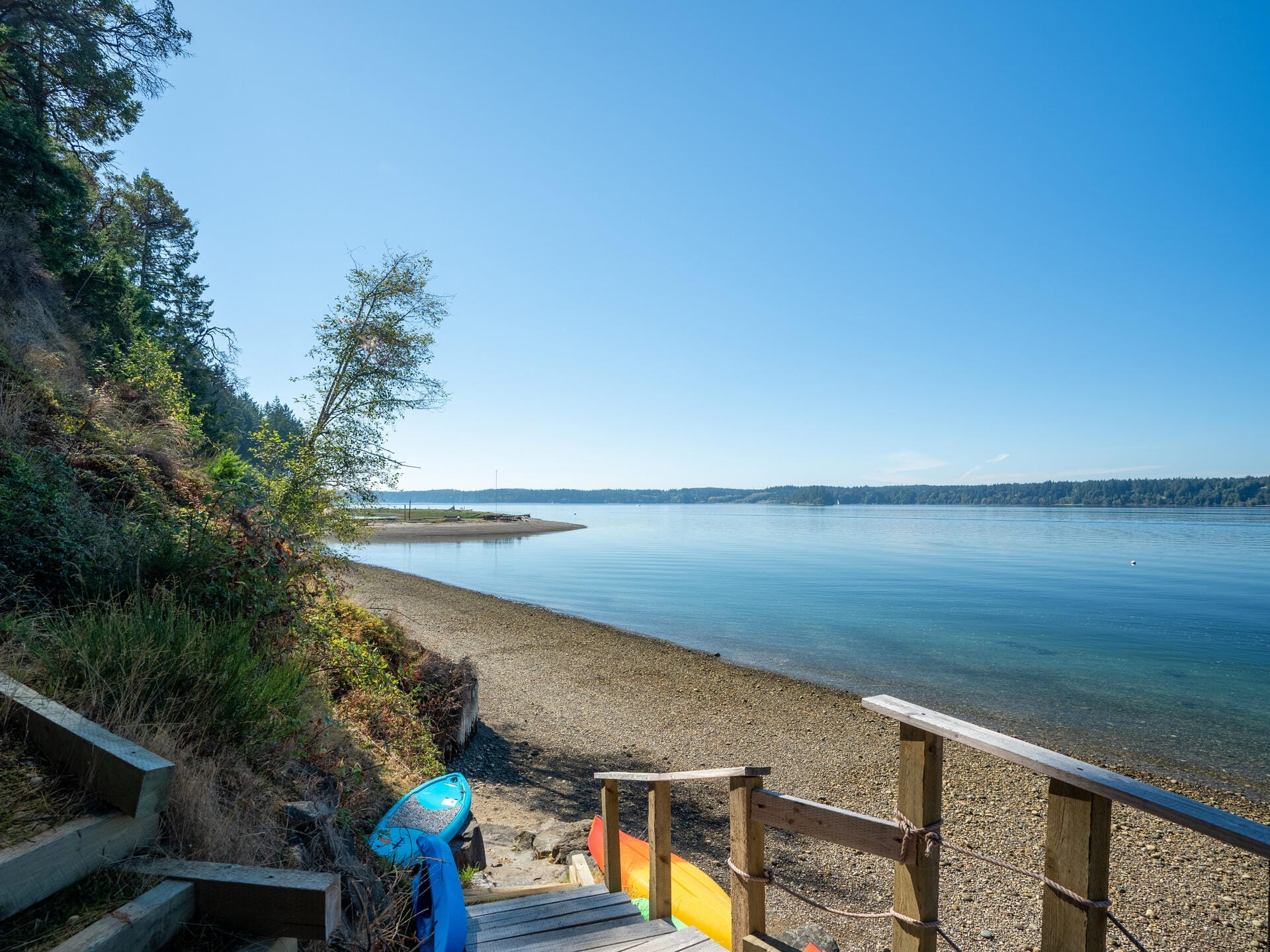 A serene beach scene with wooden stairs leading down to a rocky shoreline. A kayak rests nearby. The calm blue water reflects the clear sky, bordered by tree-lined banks stretching into the distance.