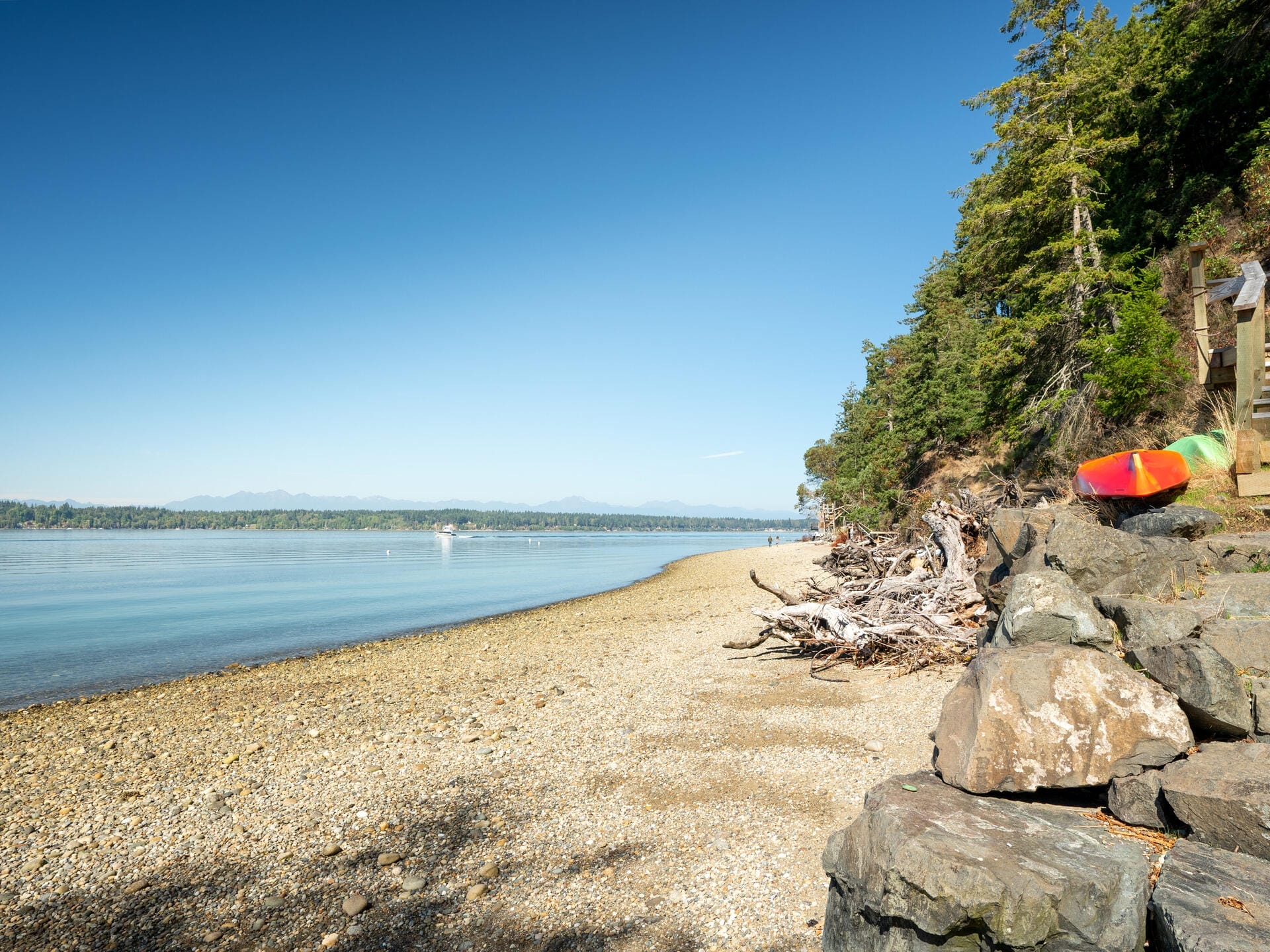 A tranquil beach scene with a rocky shore and scattered driftwood. A colorful kayak rests on a stone wall to the right. The calm water meets a distant tree-lined horizon under a clear blue sky.