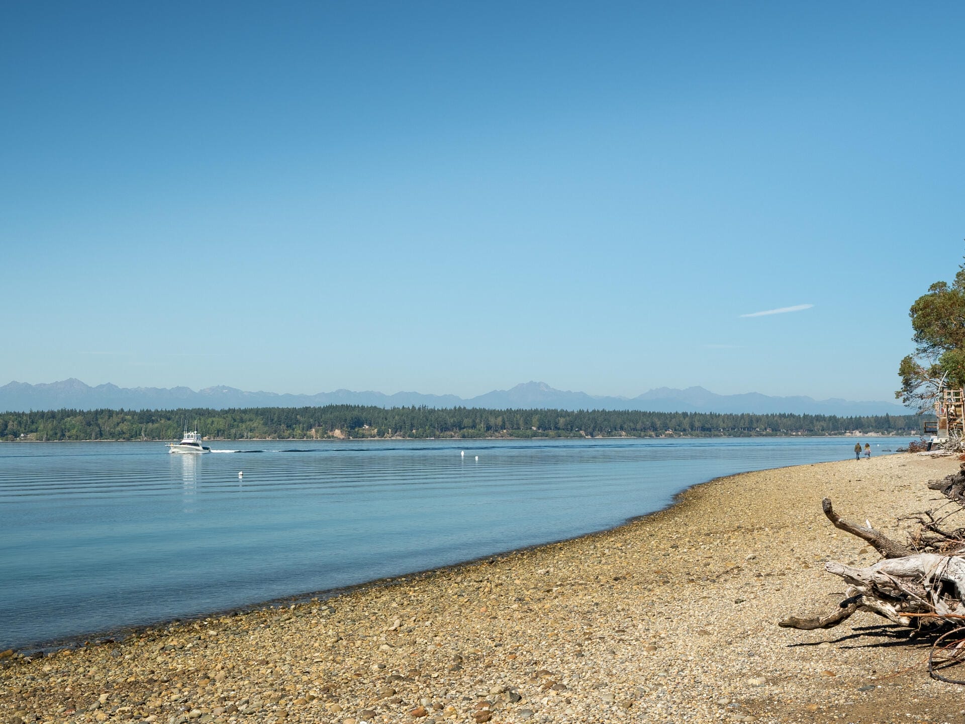 A tranquil beach scene with calm, clear blue water stretching to the horizon. A small boat is visible on the water. A rocky shoreline leads to trees on the right, under a clear sky with distant mountains in the background.