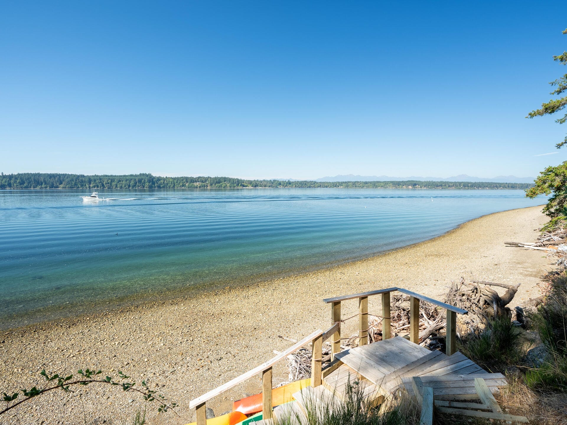 A serene view of a sandy and pebbly shoreline with calm, blue water and a clear sky. Wooden steps lead down to the beach, and a small boat is visible on the water. Forested land is seen across the bay.