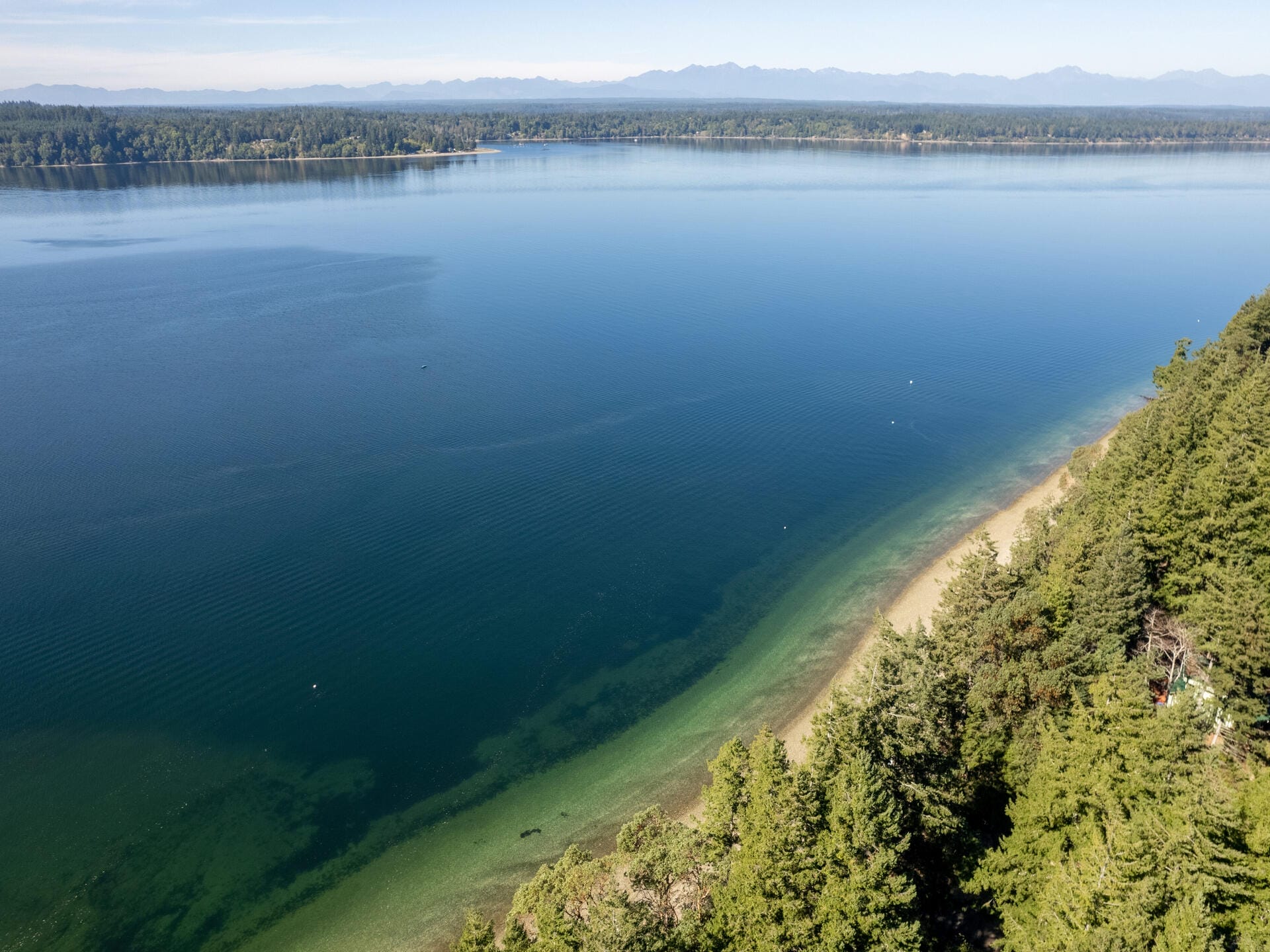 Aerial view of a serene lake with clear blue water, surrounded by dense green forests along the shoreline. Hills and mountains are visible in the background under a clear sky.