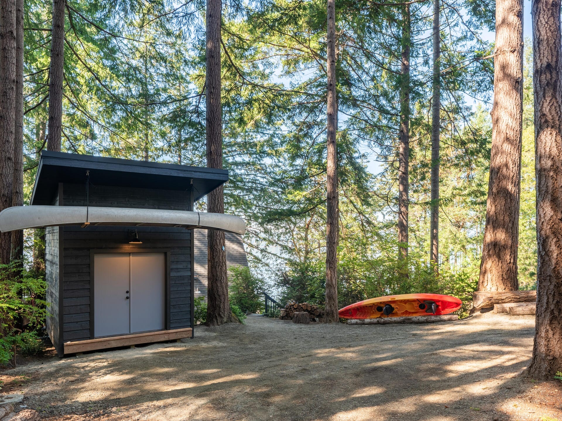 A small cabin with a canoe mounted on its side stands amidst tall trees. A red and orange kayak rests on the ground nearby. Sunlight filters through the forest, creating a peaceful, natural setting.