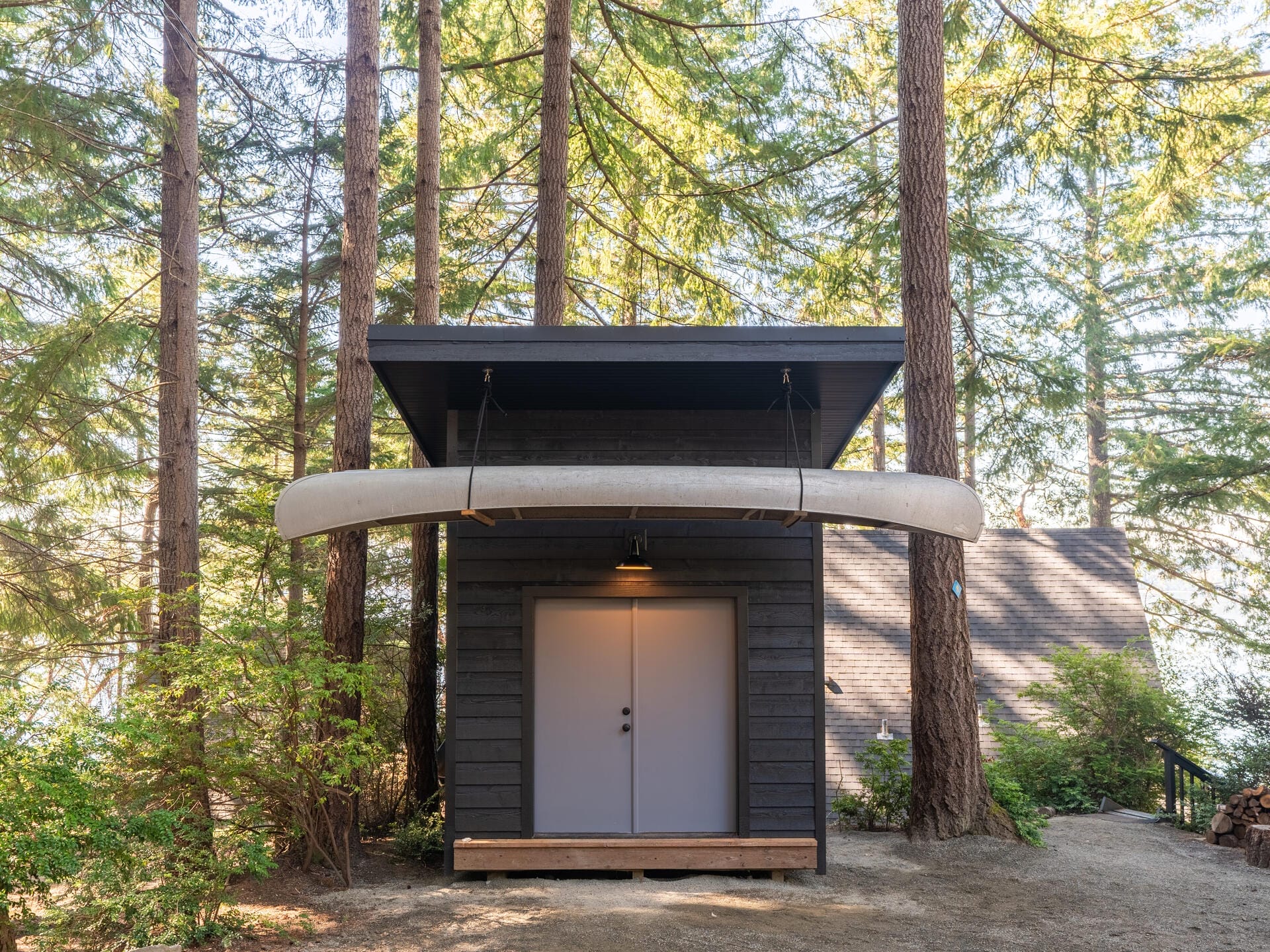 Small dark wooden cabin in a forest setting with tall trees. A canoe is mounted above the entrance. A larger house is partially visible in the background. The scene is serene and surrounded by greenery.