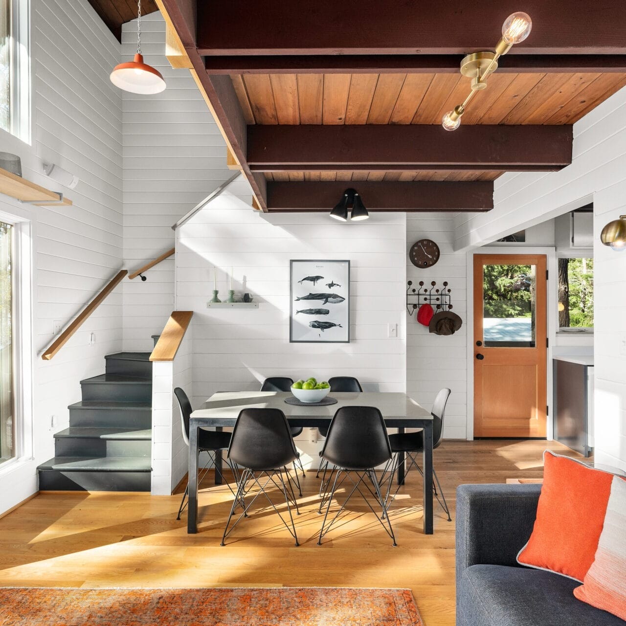 A modern living and dining area in a cozy Portland, Oregon home. Features include wooden beams, a staircase, large windows with a forest view, and a dining table with black chairs. Colorful cushions adorn the gray sofa, complemented by various wall art and decor pieces.