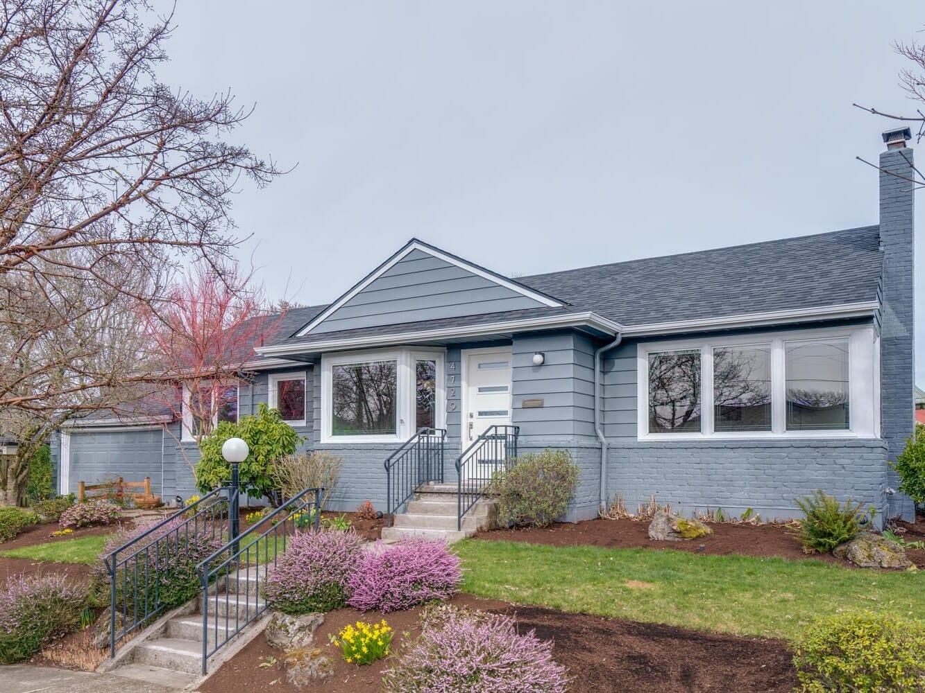 A single-story, blue-gray house in Portland, Oregon, boasts a gabled roof and white trim. The front yard features green shrubs, purple flowers, and a small leafless tree. Steps lead to the front door, flanked by metal railings.