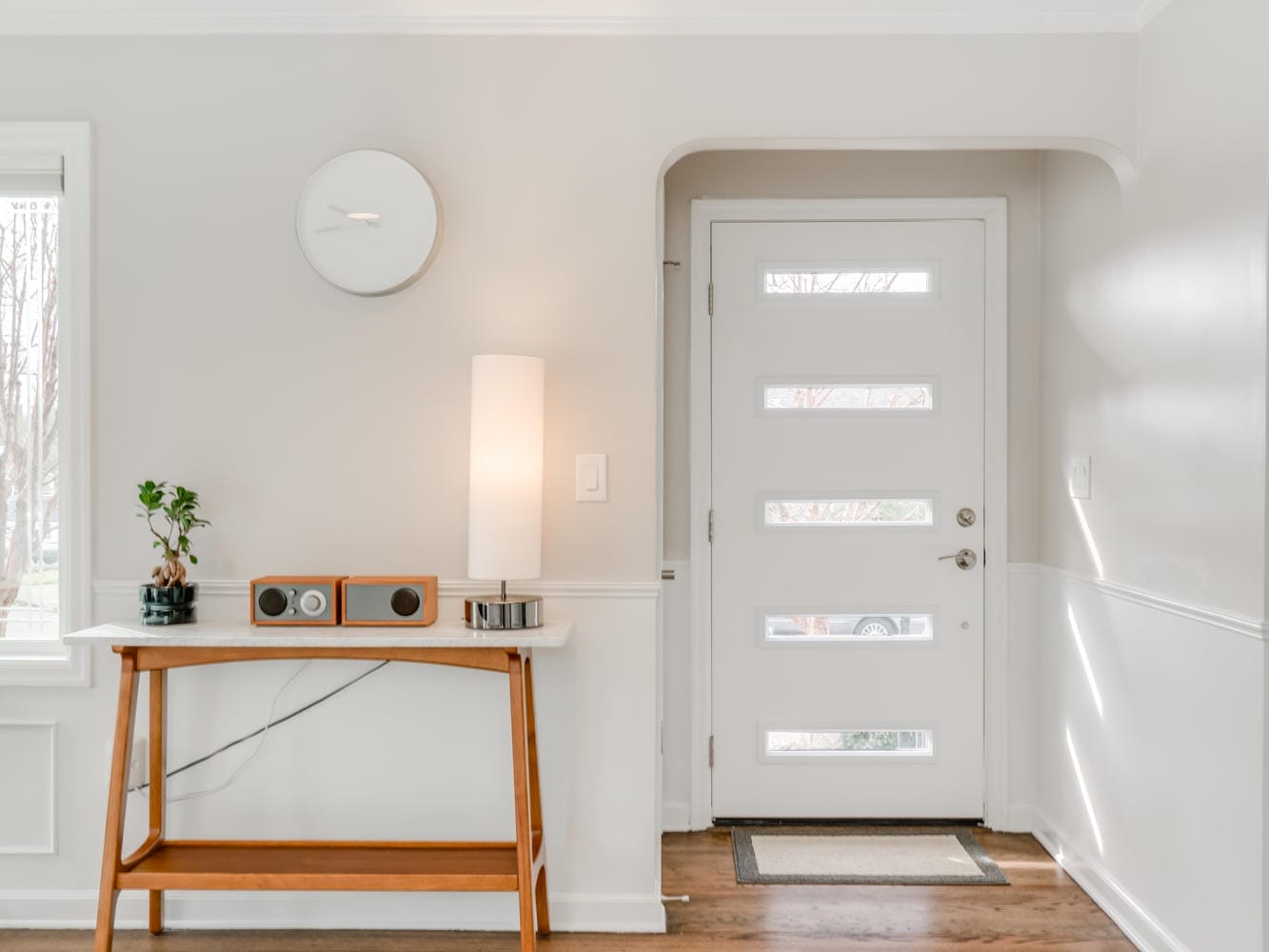 A bright, minimalist entryway in Portland, Oregon features a white door with rectangular glass panels, a wooden console table with a lamp, a plant, and a radio on top. A small round mirror hangs on the light-colored wall above the table.