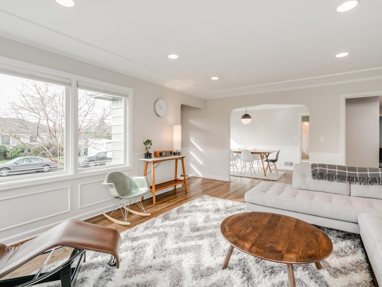 A bright, modern living room in the heart of Portland, Oregon, boasts a large window, a gray sectional sofa, a wooden coffee table, and a shag rug. A lounge chair graces the foreground while a dining area is visible through an arched opening in the background.