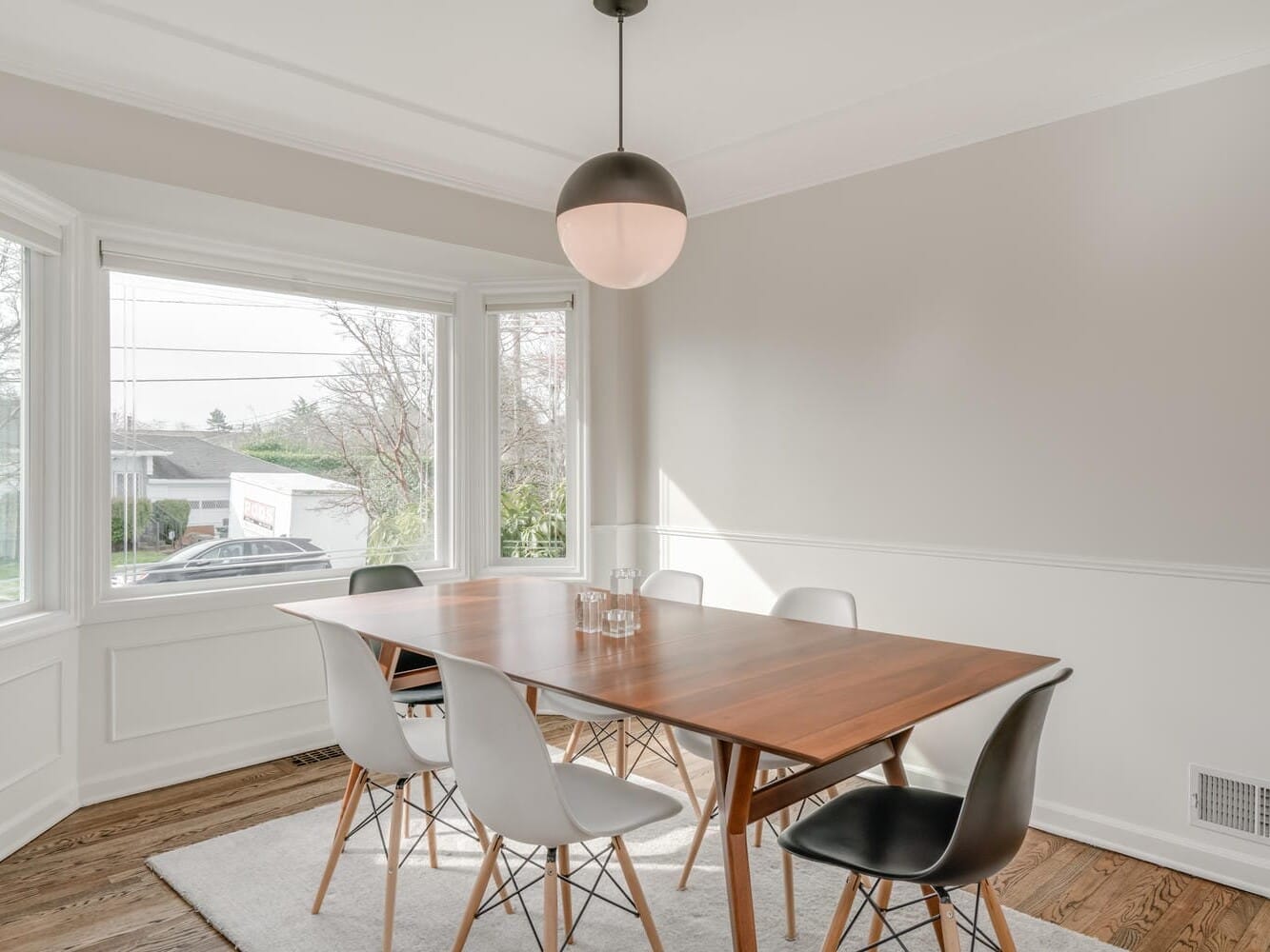 A minimalist dining room in Portland, Oregon, features a wooden table surrounded by black and white chairs on a light rug. A large window offers natural light, while a modern round pendant light hangs from the ceiling. The walls are painted in soothing light gray.