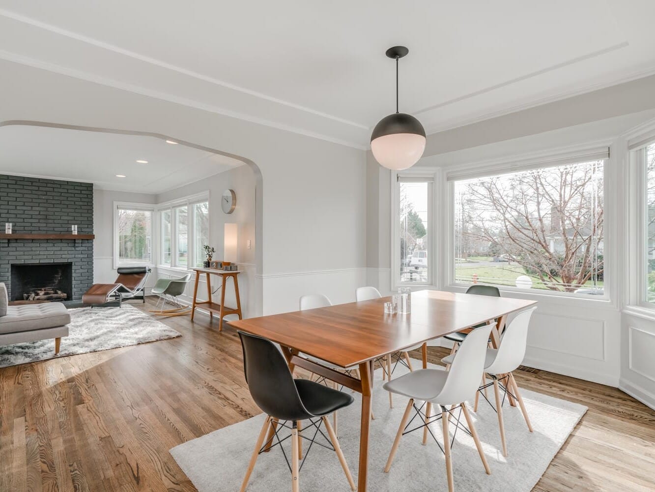 A bright dining room with a wooden table and six chairs sits in this Portland, Oregon home, featuring a large window overlooking a garden. An open archway leads to a cozy living area with a fireplace and sofa. The wooden floor complements the modern pendant light above.