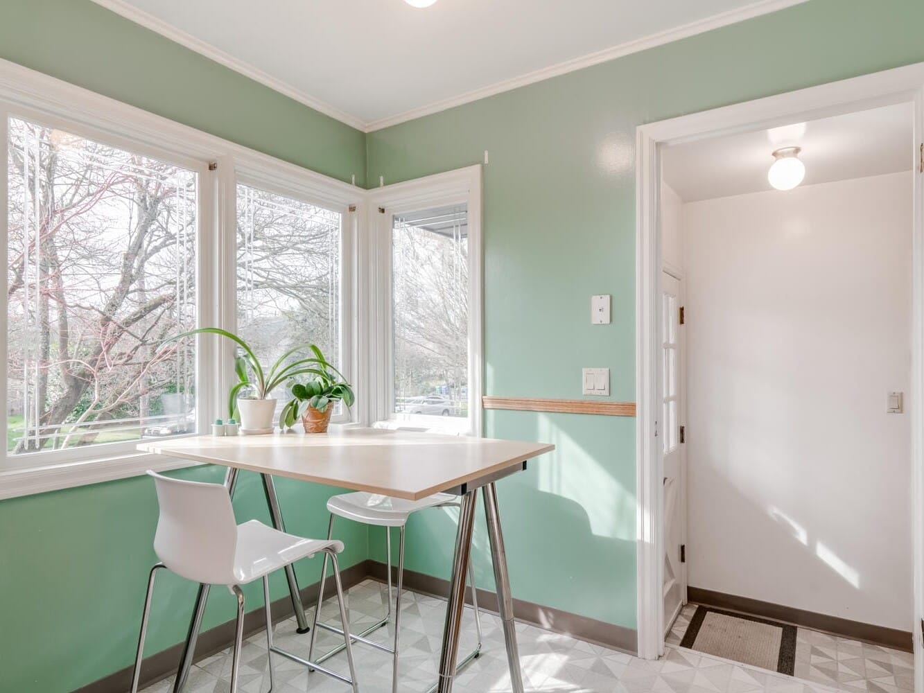 A bright kitchen corner with mint green walls features two white chairs at a high table, capturing the charm of Portland, Oregon. Large windows offer a view of leafless trees. A hallway with a door and light is visible to the right, while potted plants decorate the table.