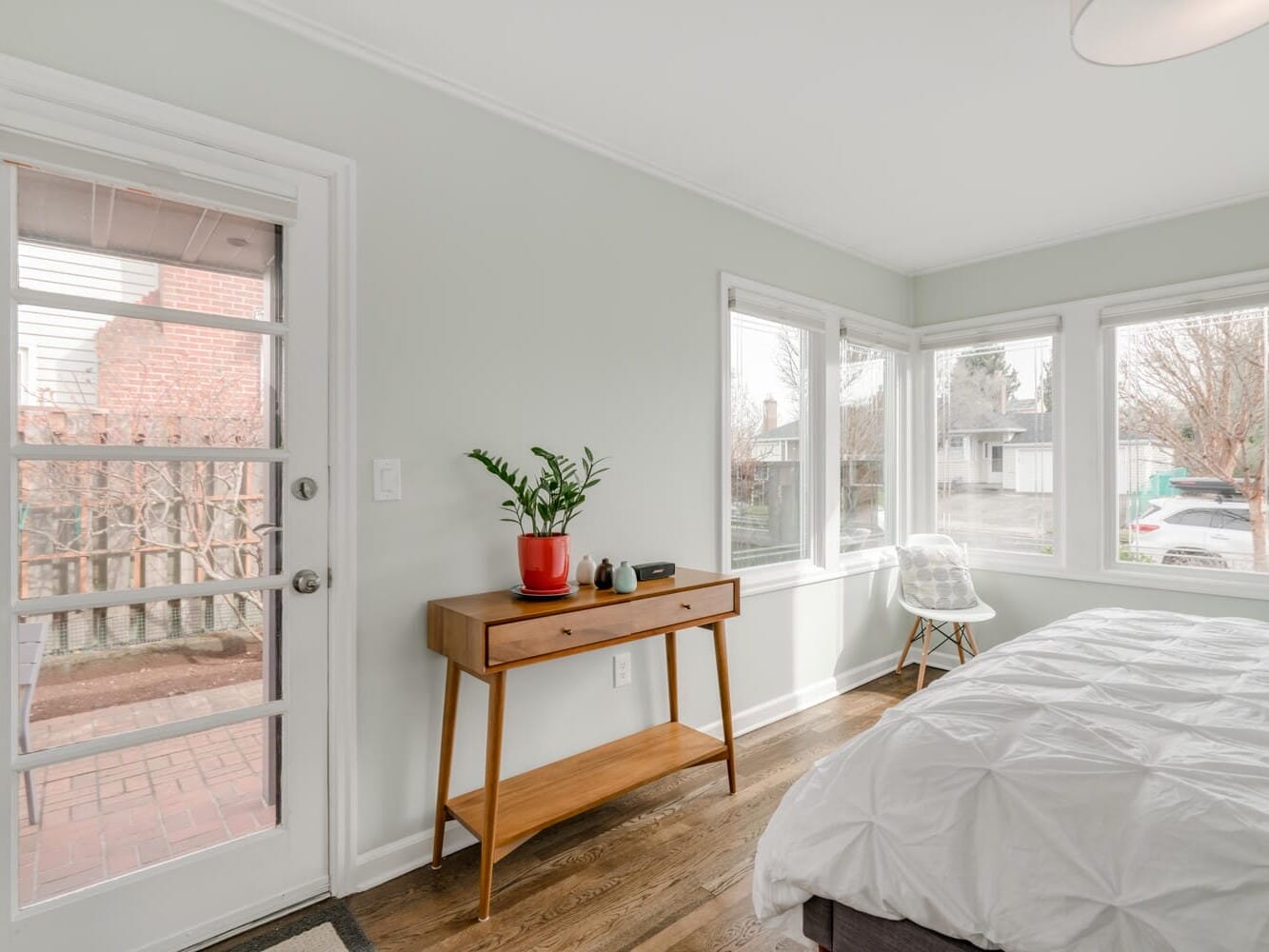 A bright bedroom in Portland, Oregon features a white bed, a wooden console table adorned with plants, and a transparent chair. Large windows reveal a tree-lined street with parked cars. A glass door opens onto a brick patio, blending indoor comfort with the city's charm.