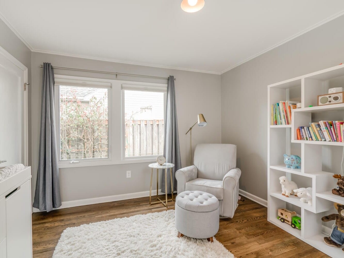 A cozy nursery in Portland, Oregon, features a white bookshelf filled with toys and books, a plush armchair with an ottoman, and a small side table. A soft rug covers the hardwood floor. Large windows with gray curtains let in natural light.