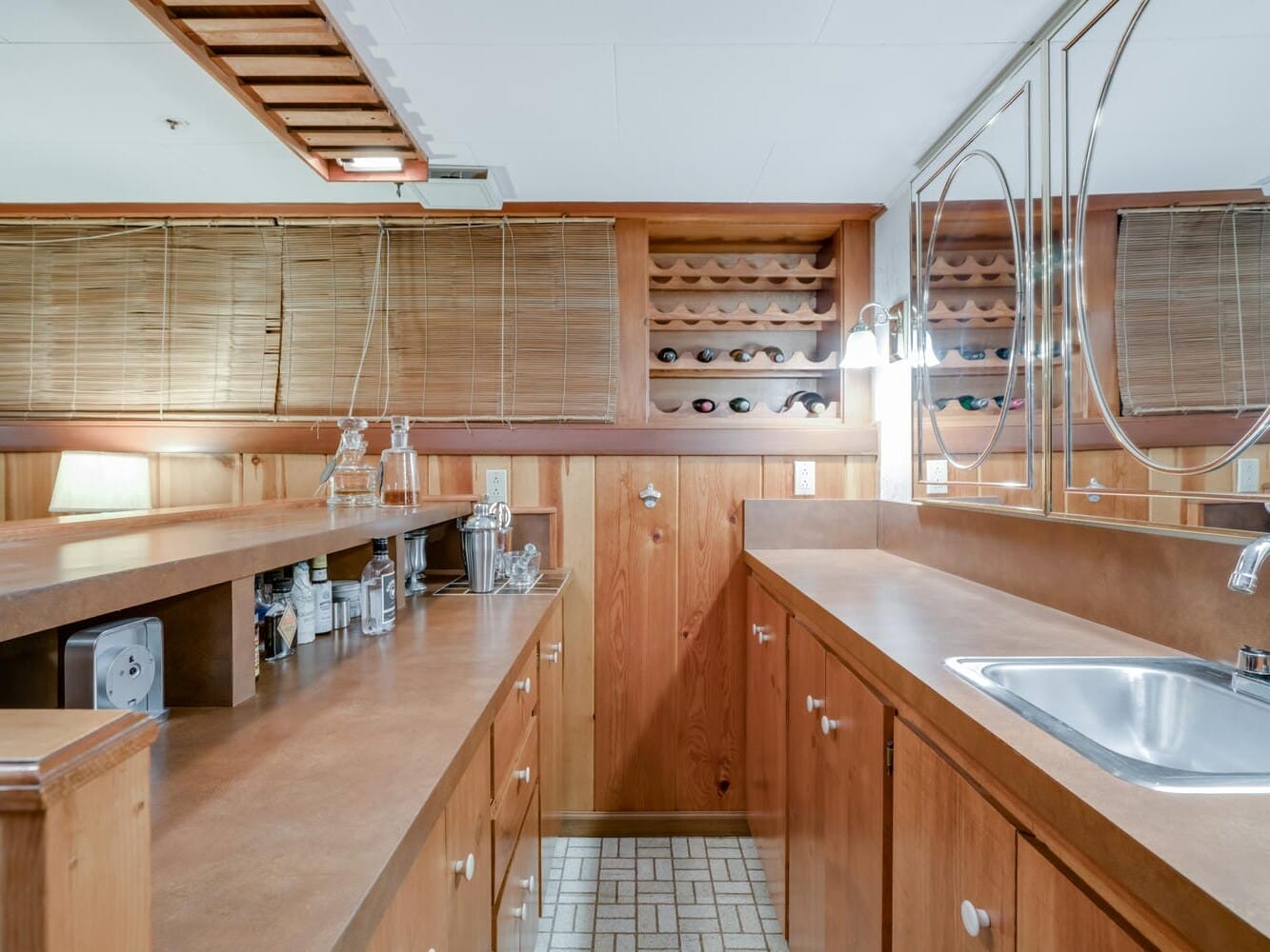 A cozy Portland, Oregon home bar features wooden cabinetry and a countertop. Several shelves hold glassware and wine bottles. A sink is embedded in the counter, while mirrors with circular designs decorate the walls. Soft lighting completes the inviting ambiance.