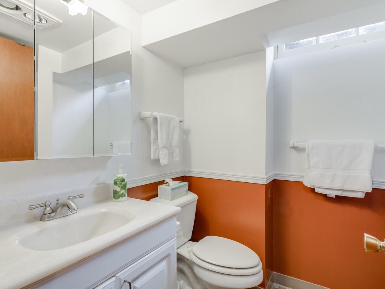 A small bathroom in Portland, Oregon, boasts orange and white walls. It features a white vanity with a sink and faucet, a mirror cabinet above, a toilet, and a window for natural light. Towels hang on the wall, while a soap dispenser and tissue box sit on the counter.