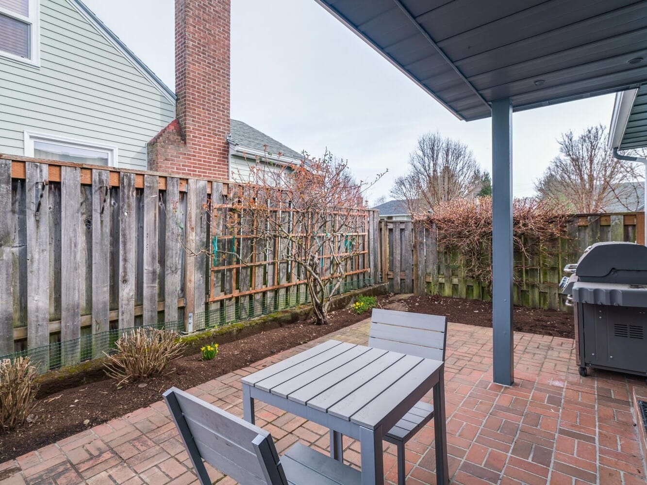 A small brick patio with a simple wooden table and two chairs, next to a barbecue grill, evokes the cozy charm of Portland, Oregon. The area, enclosed by a wooden fence, is framed by bare trees and shrubs along the edges, with houses visible in the background.