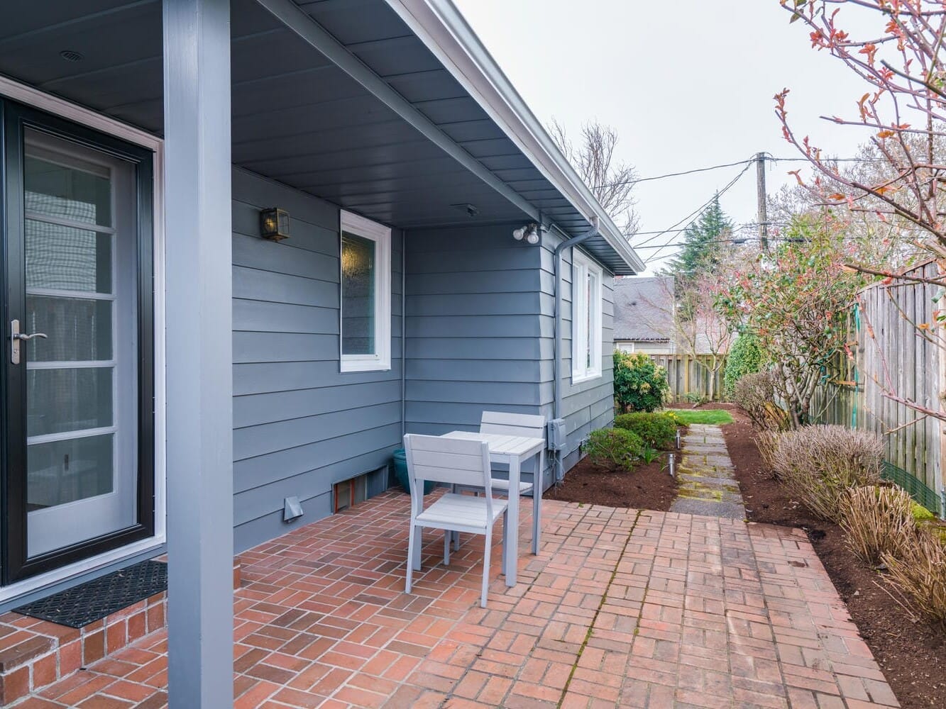 A small patio with a brick floor features a table and two chairs, perfectly capturing the charm of Portland, Oregon. The house exterior is painted blue-gray with a glass door. Nearby, a garden with shrubs and a tree is bordered by a wooden fence on this overcast day.