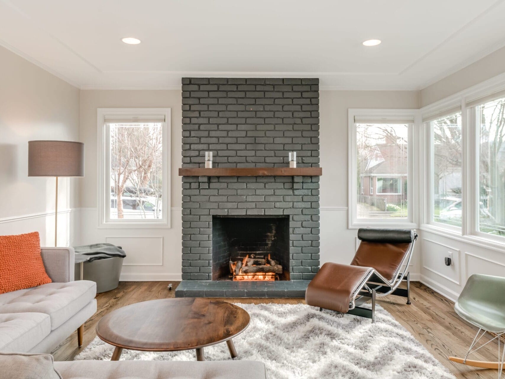 A cozy Portland, Oregon living room features a gray brick fireplace, modern furnishings, and wooden floor. A beige sofa sits beside a round wooden table and a chair. Large windows let natural light fill the space, while a fluffy rug adds warmth to the inviting atmosphere.