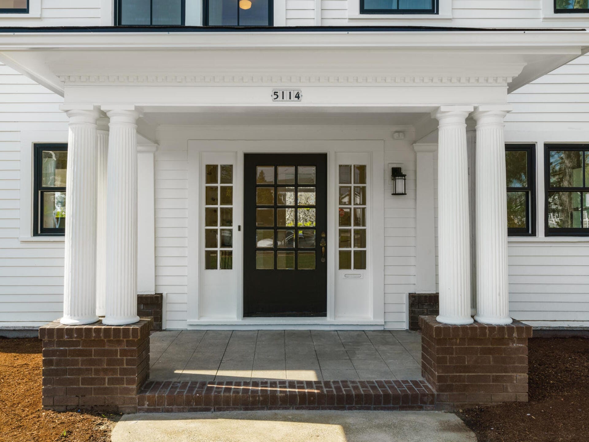 A white house entrance with four white columns and brick bases. The dark green front door has multiple glass panes, and the number 5114 is above the entrance. Theres a wall-mounted lantern on the right.