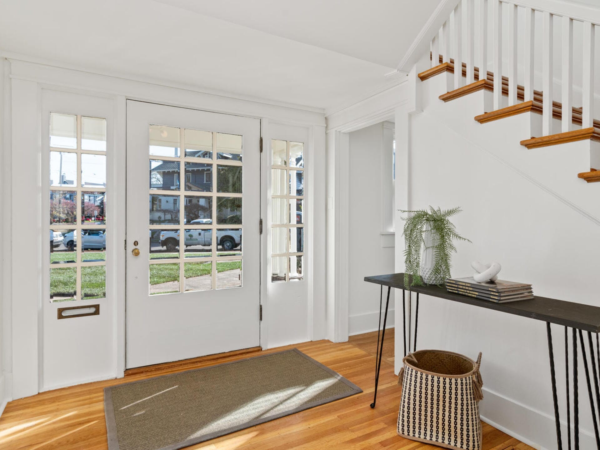 A bright entryway with a door featuring glass panels, allowing natural light to illuminate the space. A staircase with wooden steps and a railing is on the right. A black console table and wicker basket are set against the white walls.