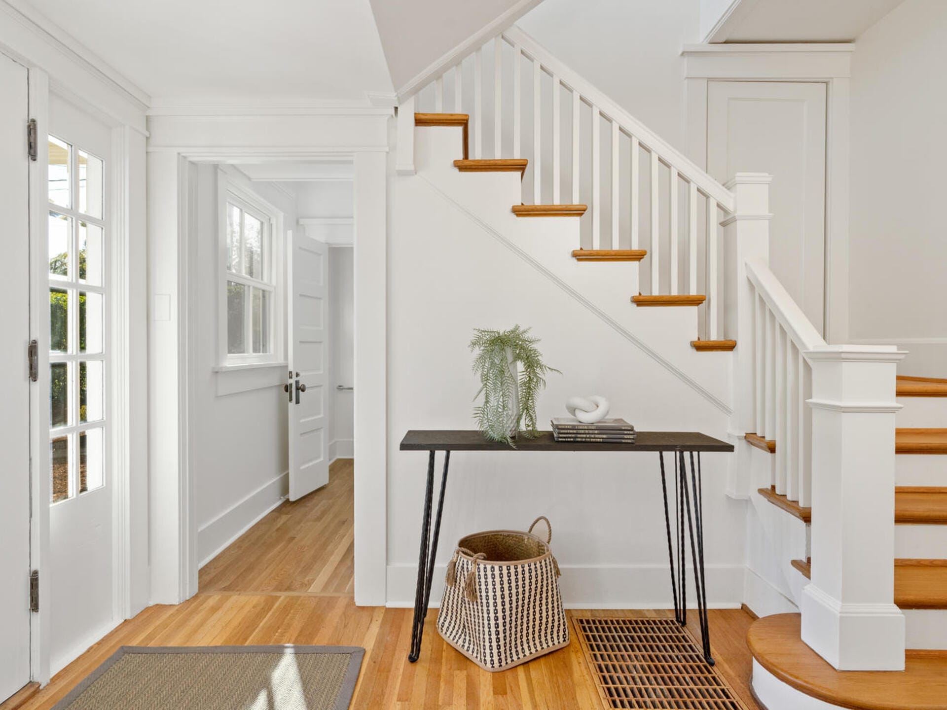 A bright entryway features a staircase with wooden steps and white railing. A narrow black console table holds books and a small plant. A basket sits on the floor nearby. Sunlight streams through a glass-paneled door, illuminating the hardwood floor.