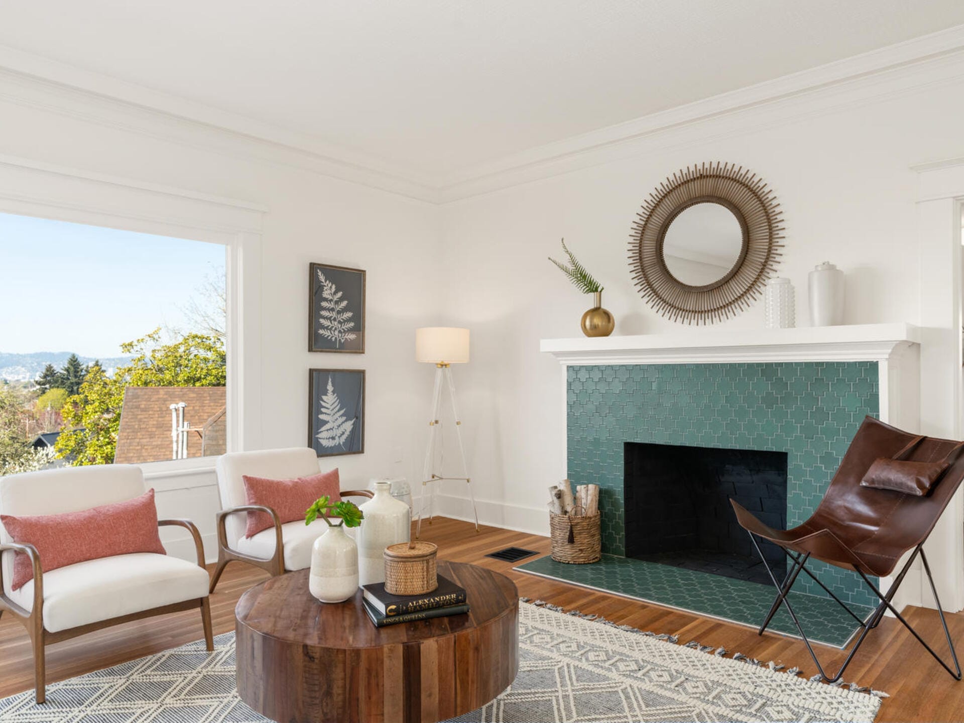 A bright living room featuring a green-tiled fireplace with a round mirror above it. The room includes white armchairs with pink cushions, a brown butterfly chair, a wooden coffee table, and a floor lamp, with a large window offering a scenic view.