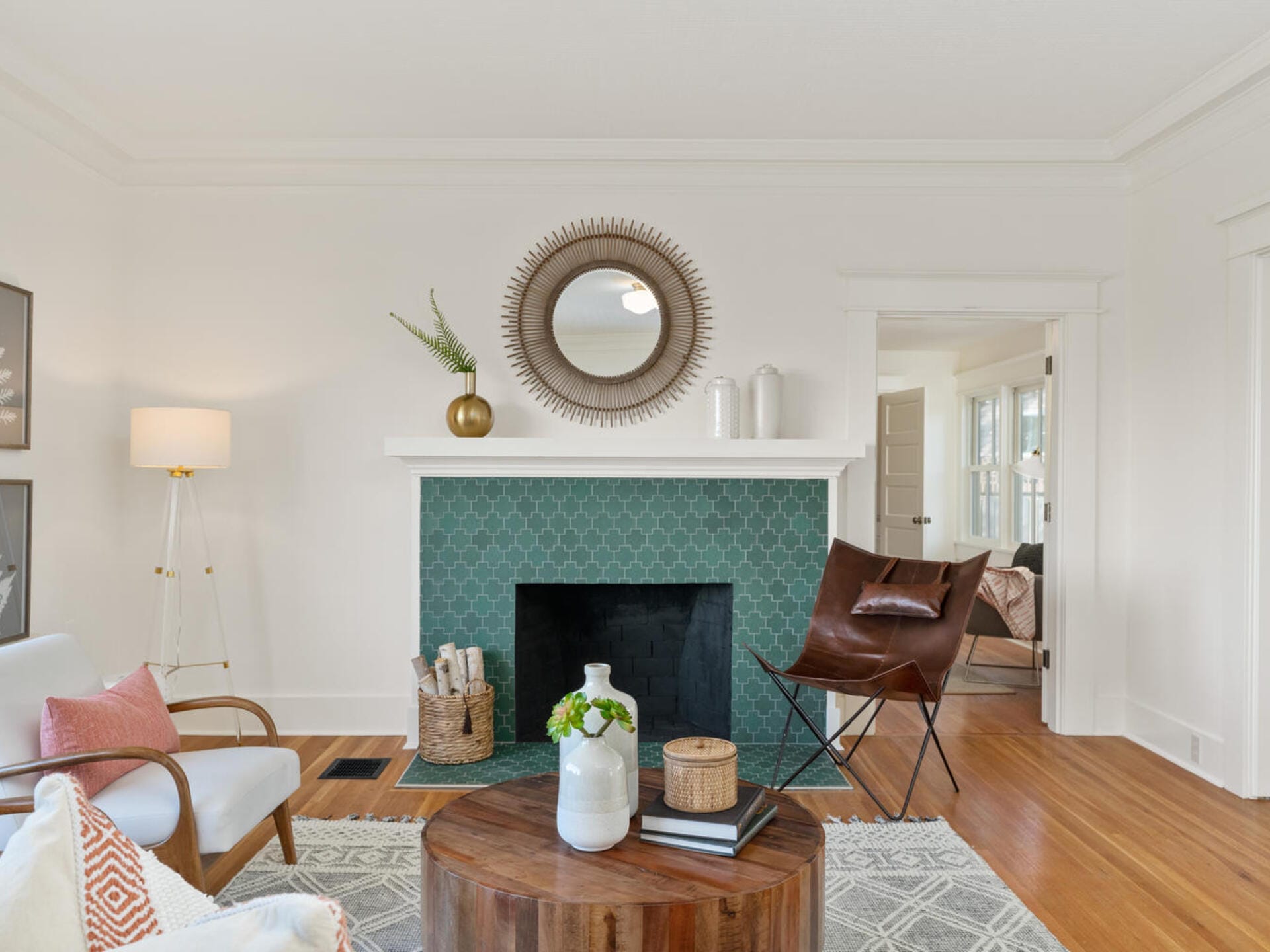 A cozy living room with a green-tiled fireplace, a sunburst mirror above, and modern decor. Features a brown leather chair, round wooden coffee table with a vase and books, a white armchair, and a white floor lamp. Light pours in from an adjacent room.