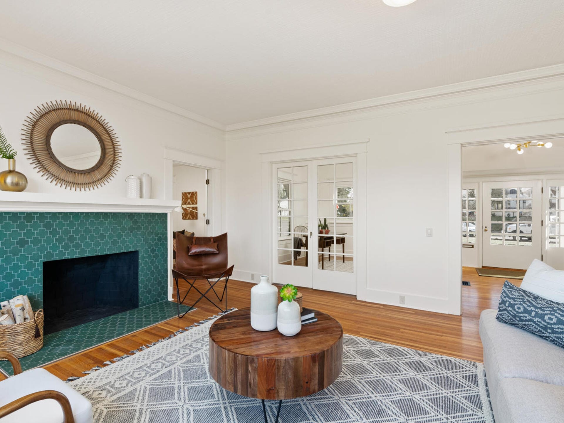A bright living room with a green tiled fireplace, round mirror, and contemporary decor. A round wooden coffee table sits on a patterned rug, with a chair and white vase accents. Double doors lead to an adjoining room with more natural light.
