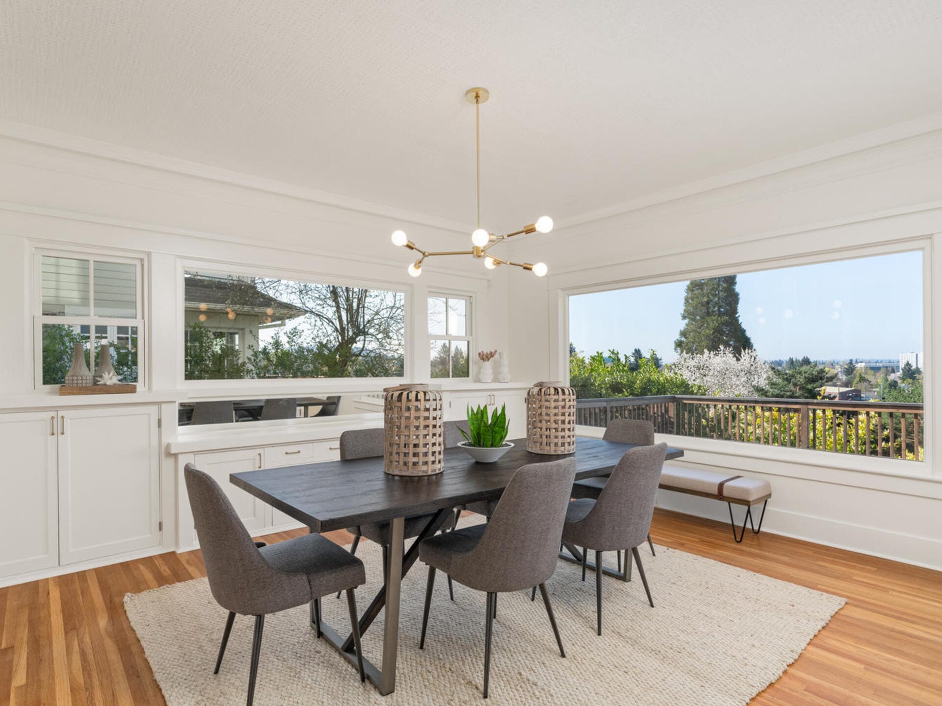 A bright dining room with a large window showcasing a scenic view. The room features a dark wooden table, gray chairs, and a modern chandelier. A beige rug covers the hardwood floor, and woven baskets serve as table decor.