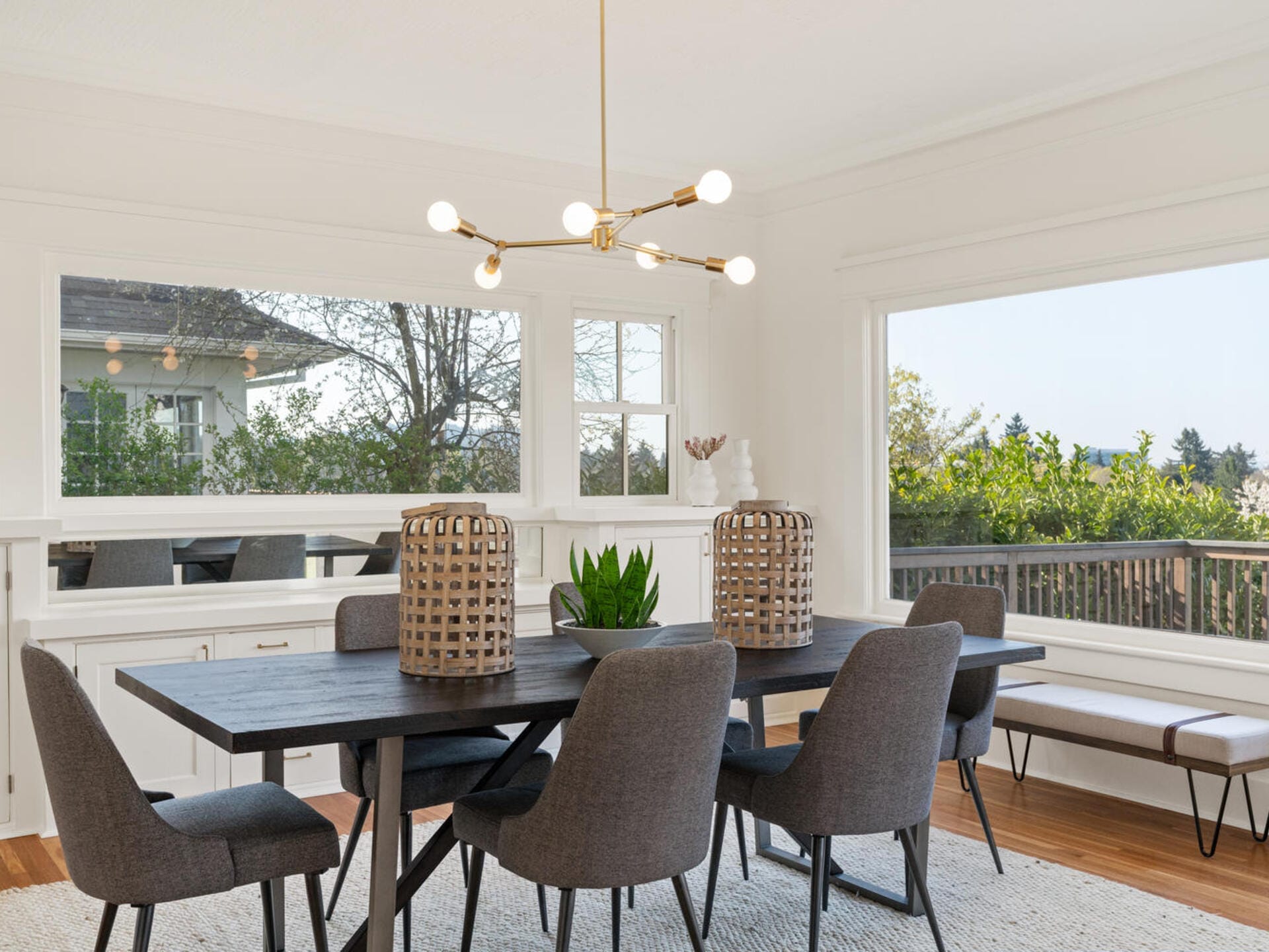 A modern dining room with a dark wooden table, six gray upholstered chairs, and a central plant centerpiece. Overhead is a gold chandelier with bulb lights. Large windows offer a view of trees and a clear sky.