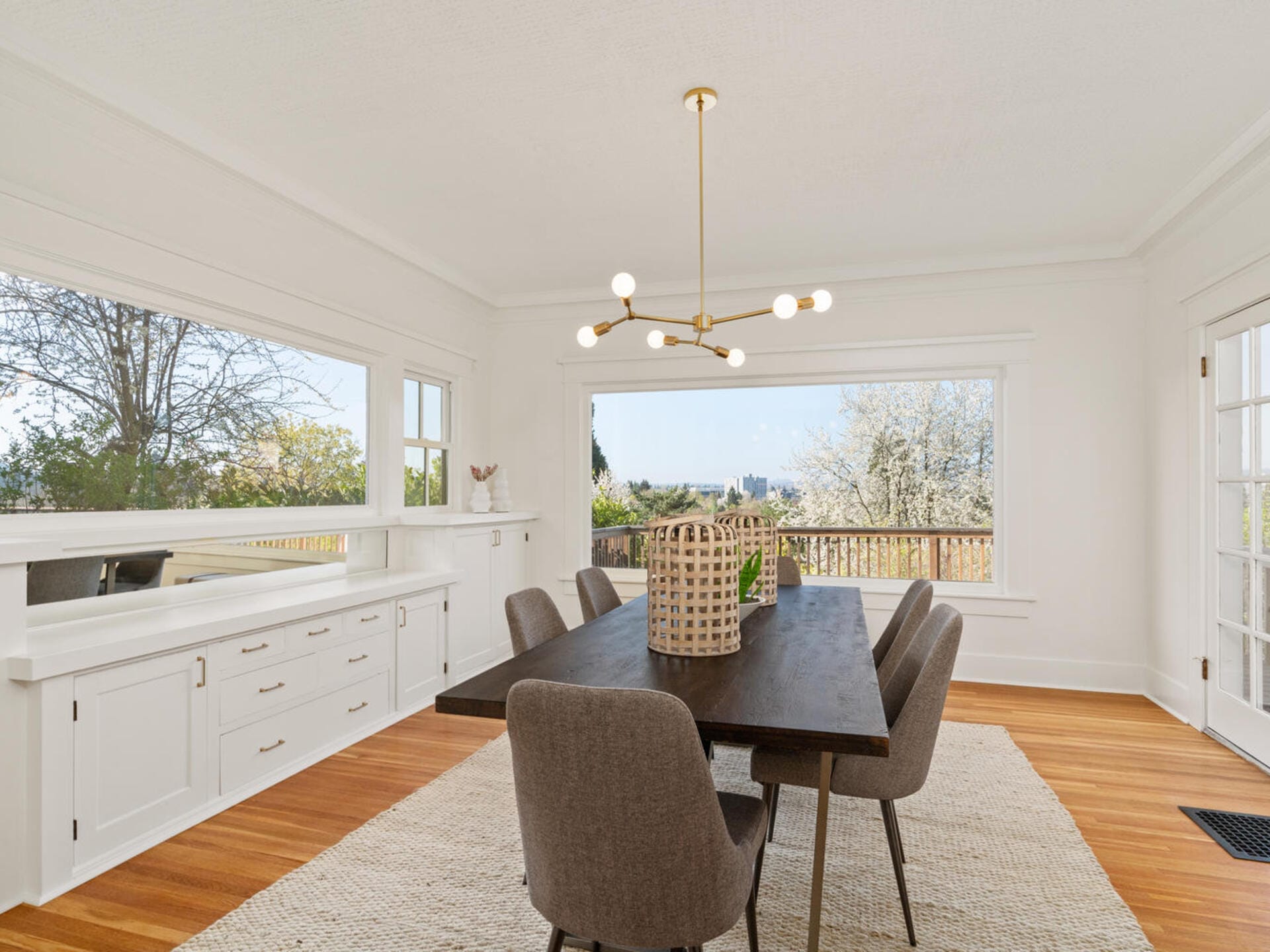 A bright dining room with a dark wood table and six gray chairs. A modern chandelier hangs overhead. Large windows offer a view of trees and a deck outside. White walls and cabinetry enhance the airy feel. A textured rug covers the hardwood floor.